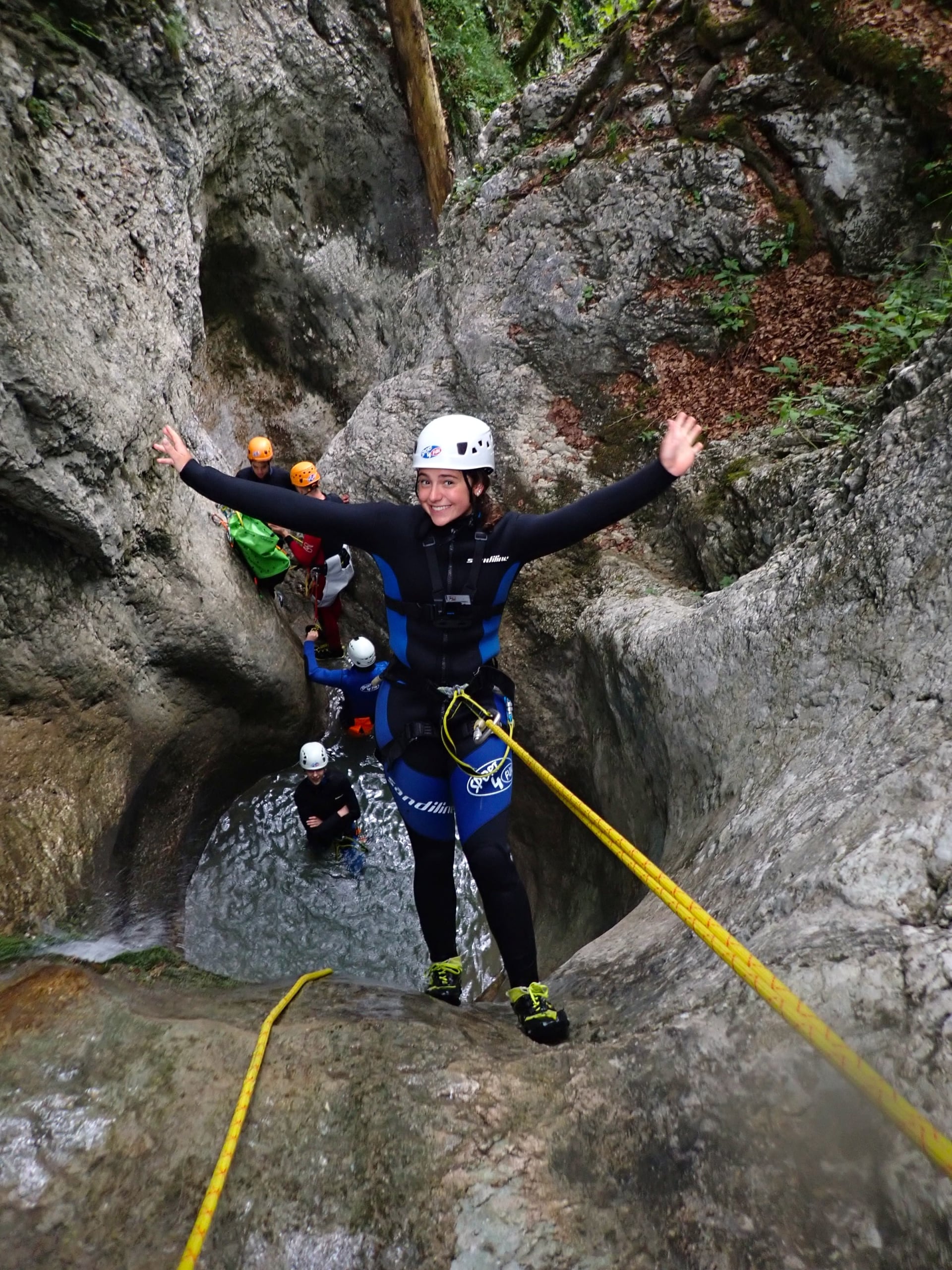 Canyoning group in a narrow rocky gorge; person in wetsuit and white helmet raises arms, teammates with helmets below, rope visible.