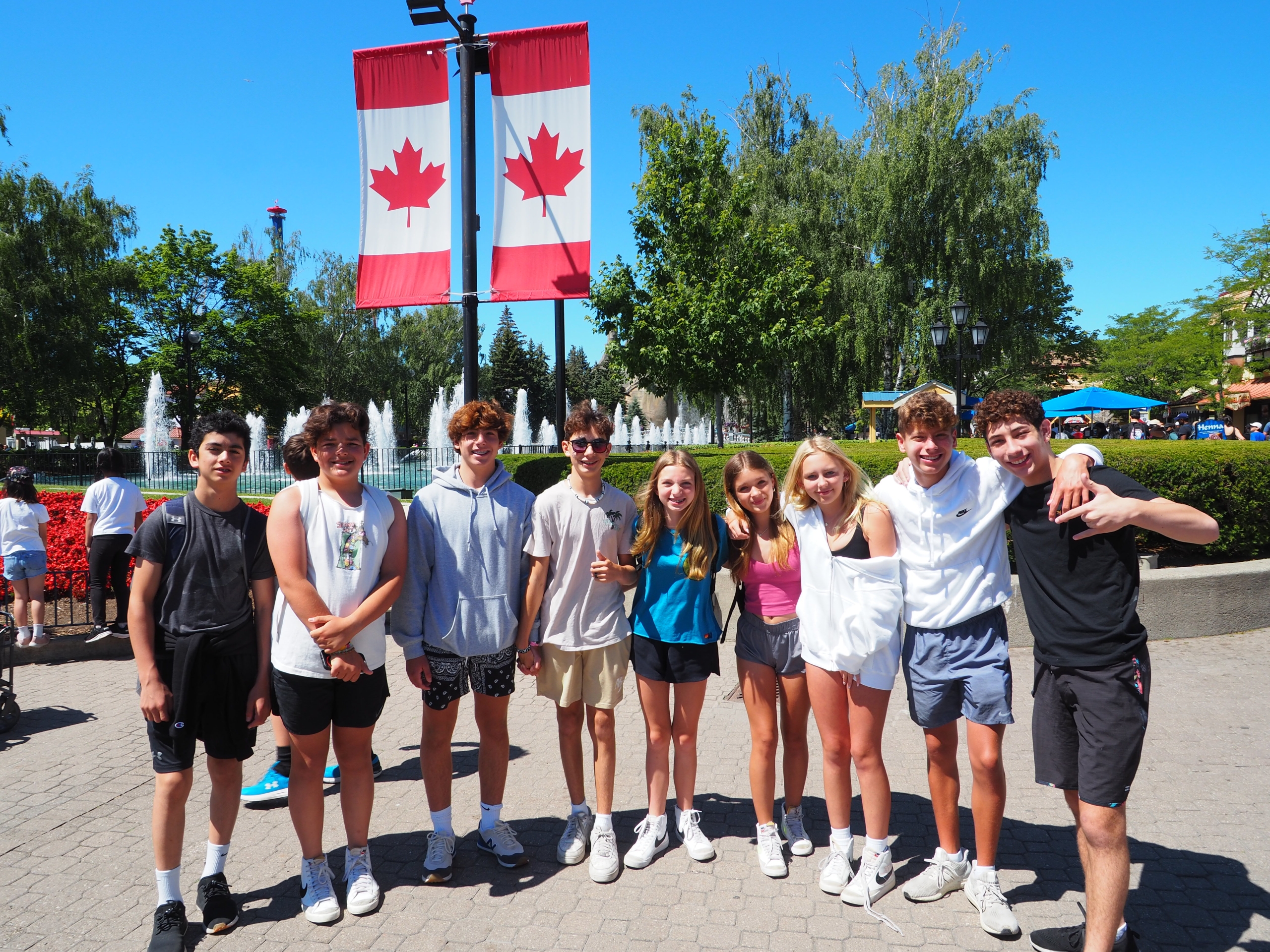 Teenagers posing together outdoors on a sunny day, with two Canadian flags on a lamppost behind them and a fountain in the background.