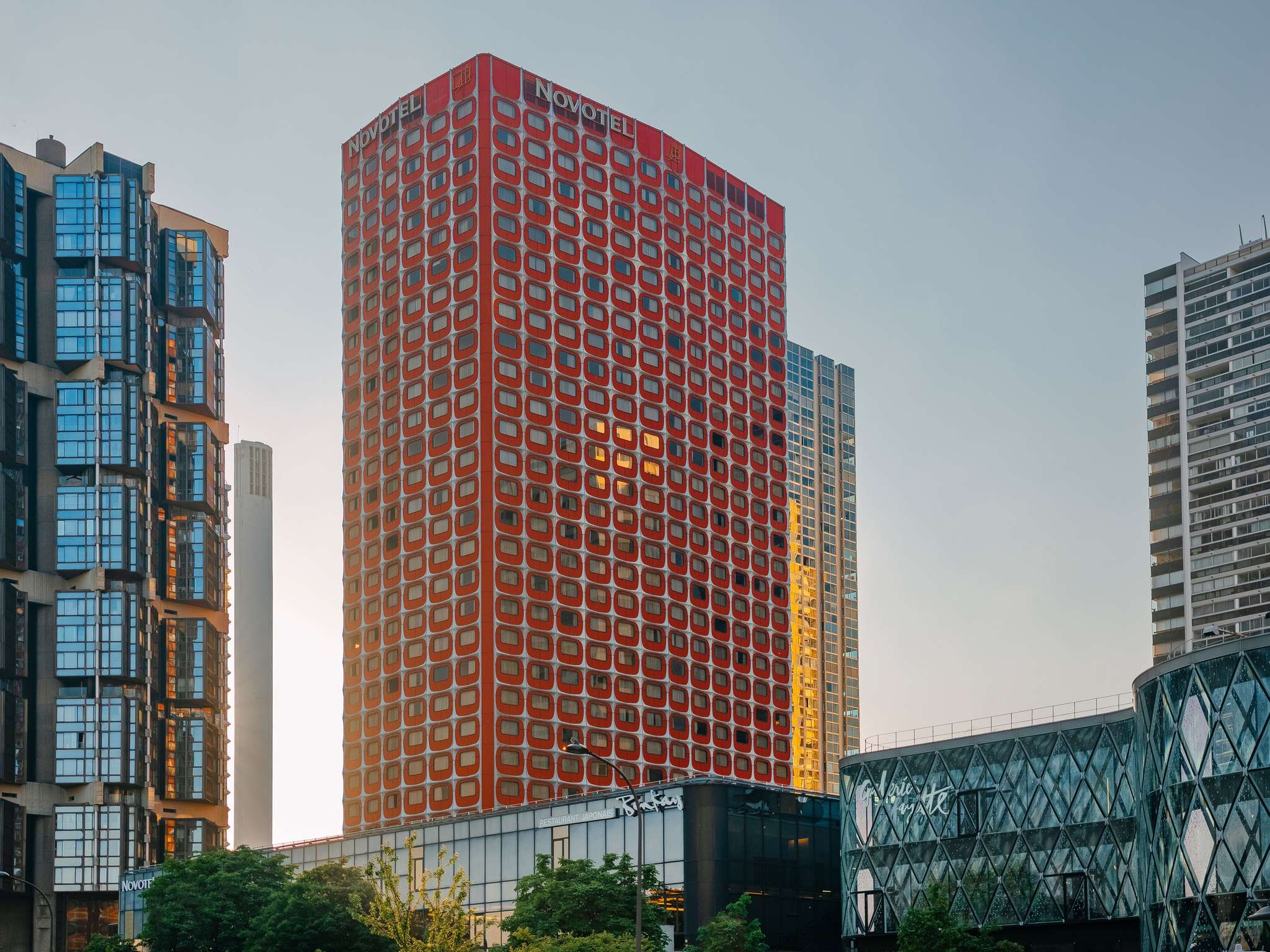 Red high-rise building with square red windows and a Novotel sign at the top, flanked by blue-glass and modern buildings under a clear sky