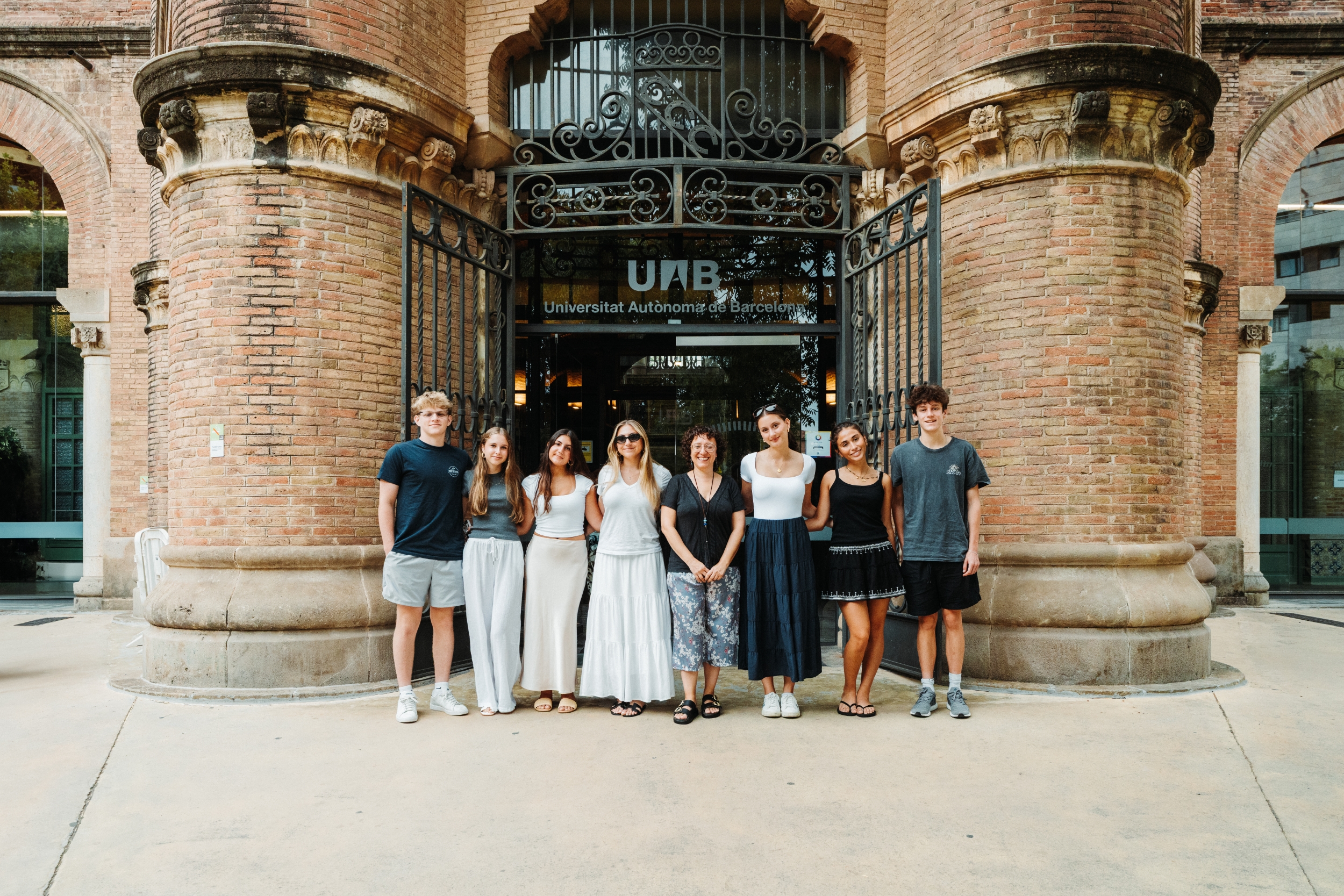 Group of eight young people standing arm-in-arm in front of a brick university building entrance with ornate iron gates (UAB sign visible).
