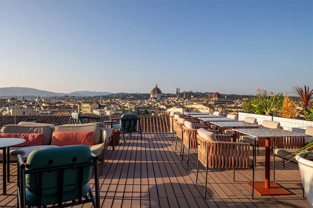 Rooftop terrace with wicker chairs and tables overlooking Florence's skyline, including the Duomo in the distance at sunset.
