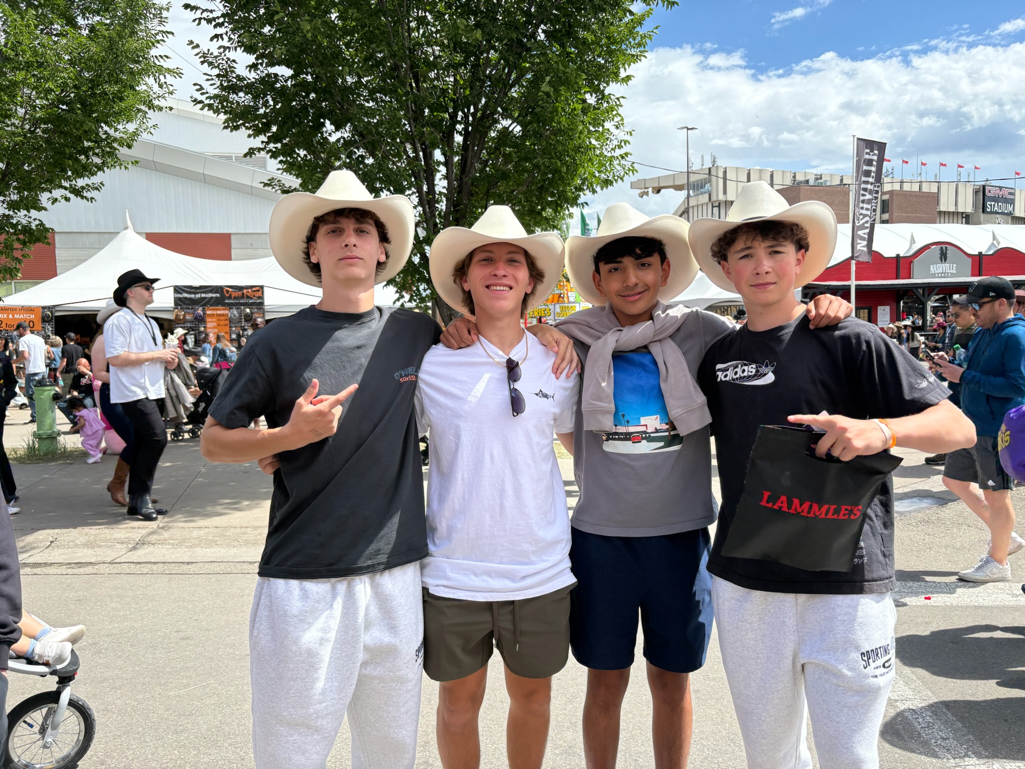 Four young men in cowboy hats pose together at an outdoor festival, smiling with arms around each other.