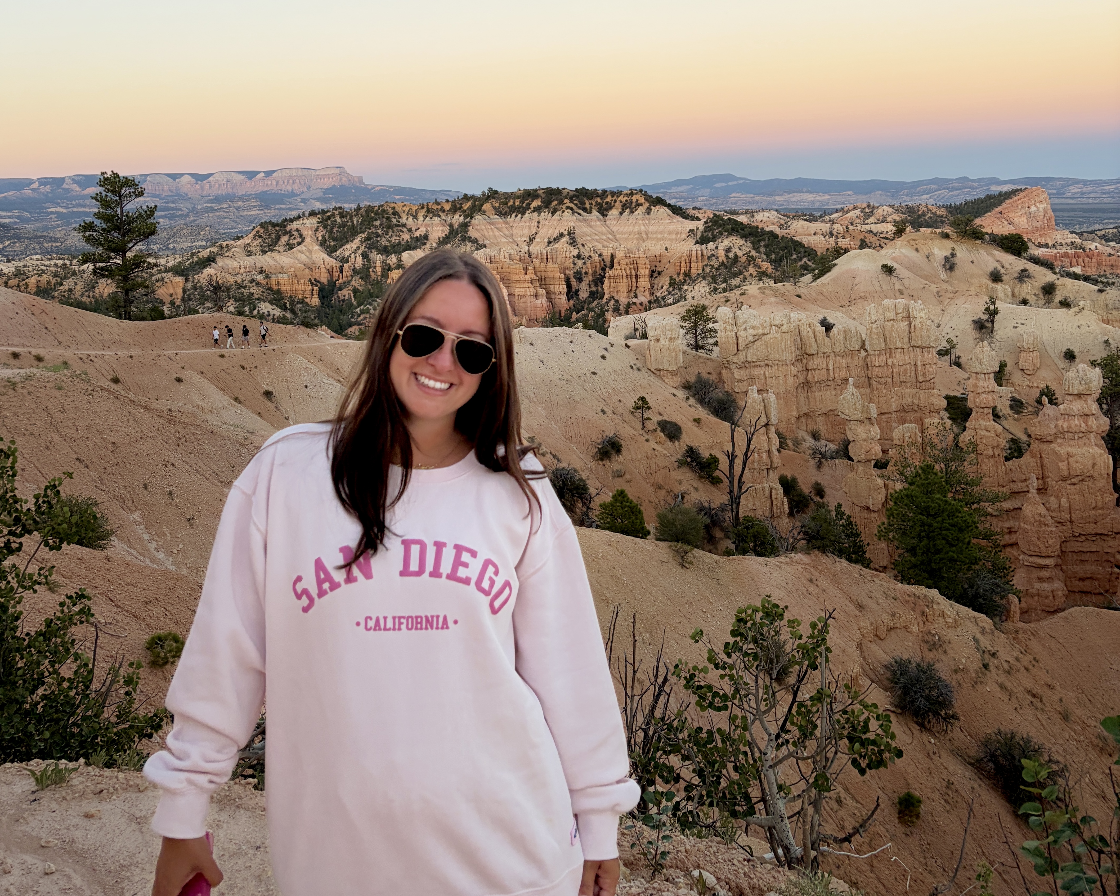 Smiling woman in a pale pink sweatshirt in the foreground with a desert canyon landscape at sunset behind her.
