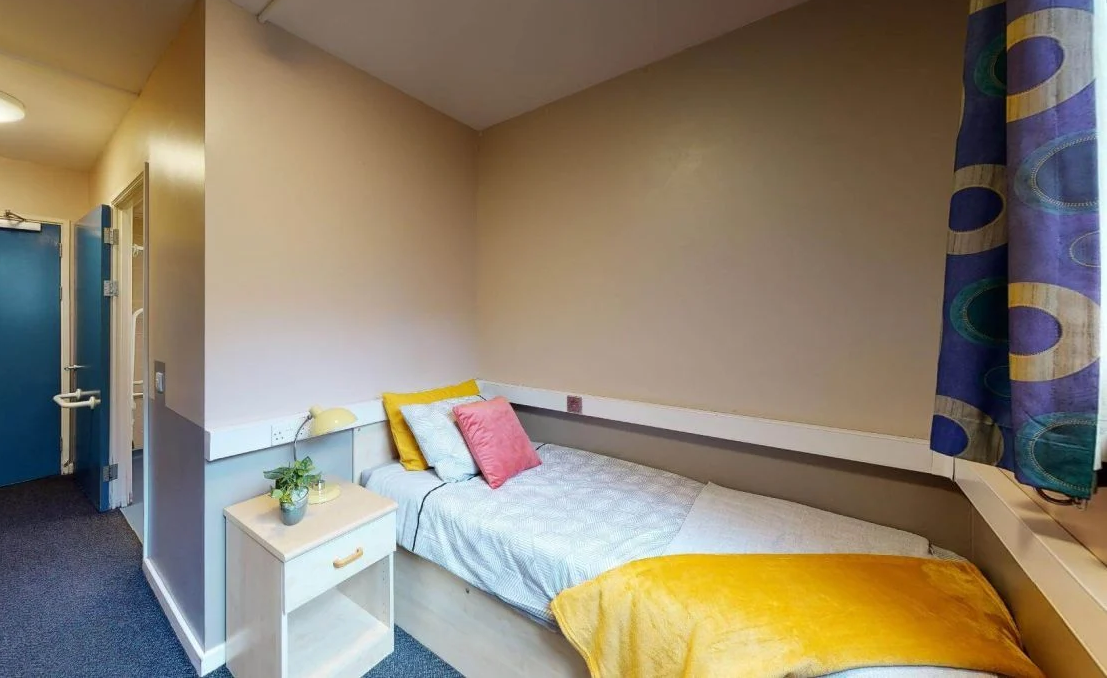 Small bedroom with a single bed, blue pillows and a pink cushion, yellow throw, white nightstand with a plant, and colorful curtains.