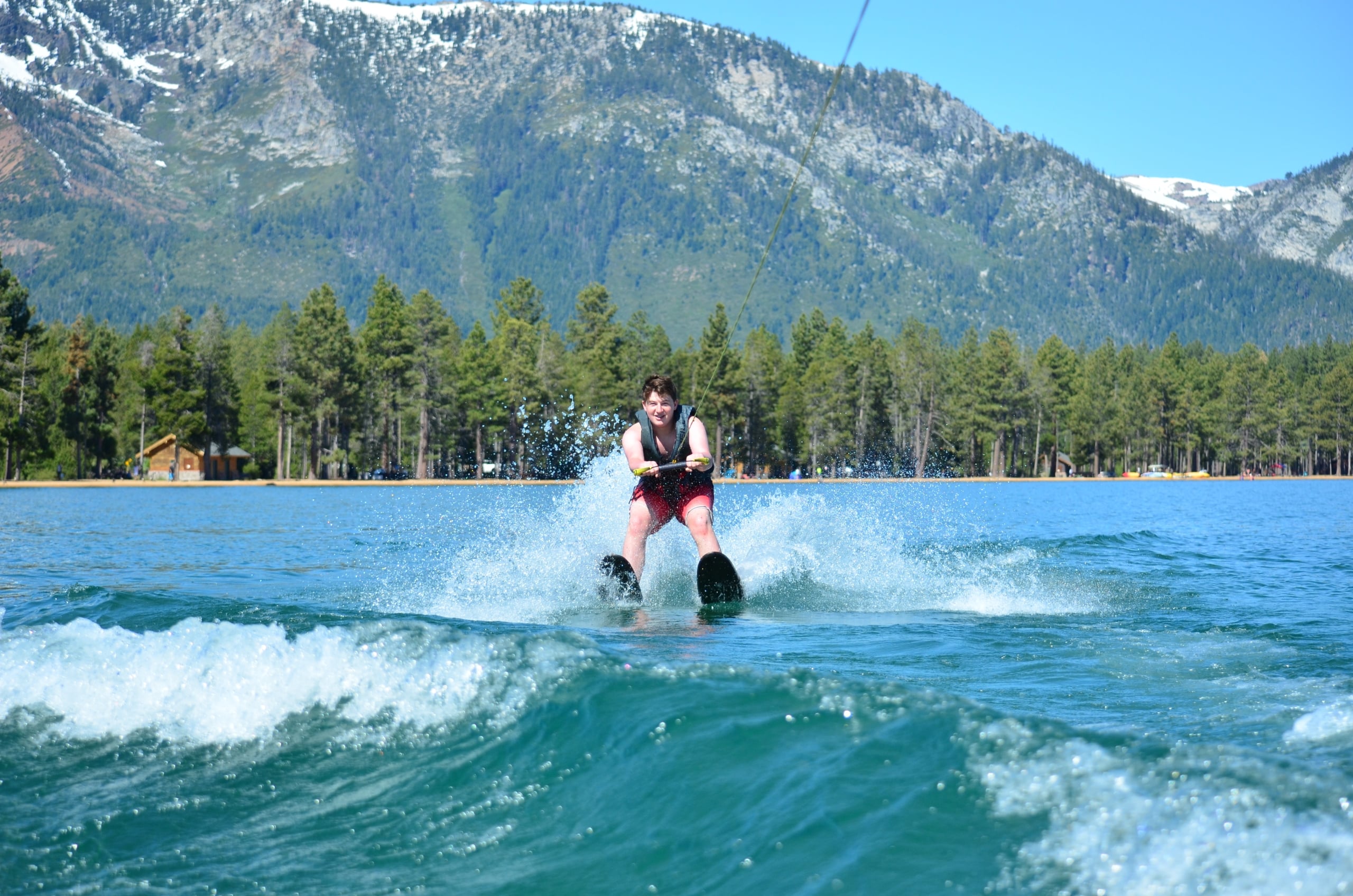 Wakeboarder on a blue lake with forested shore and snow-capped mountains in the background, wearing a life vest.