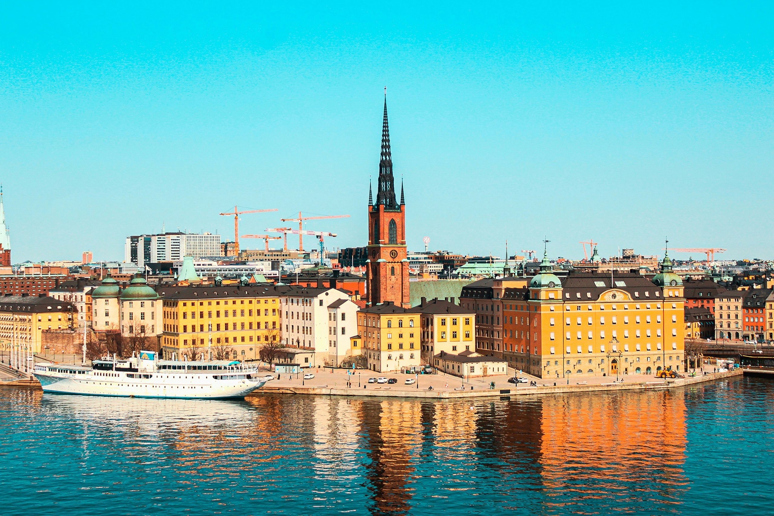 City waterfront with a tall pointed church spire rising above pastel buildings and a white boat docked in the foreground.