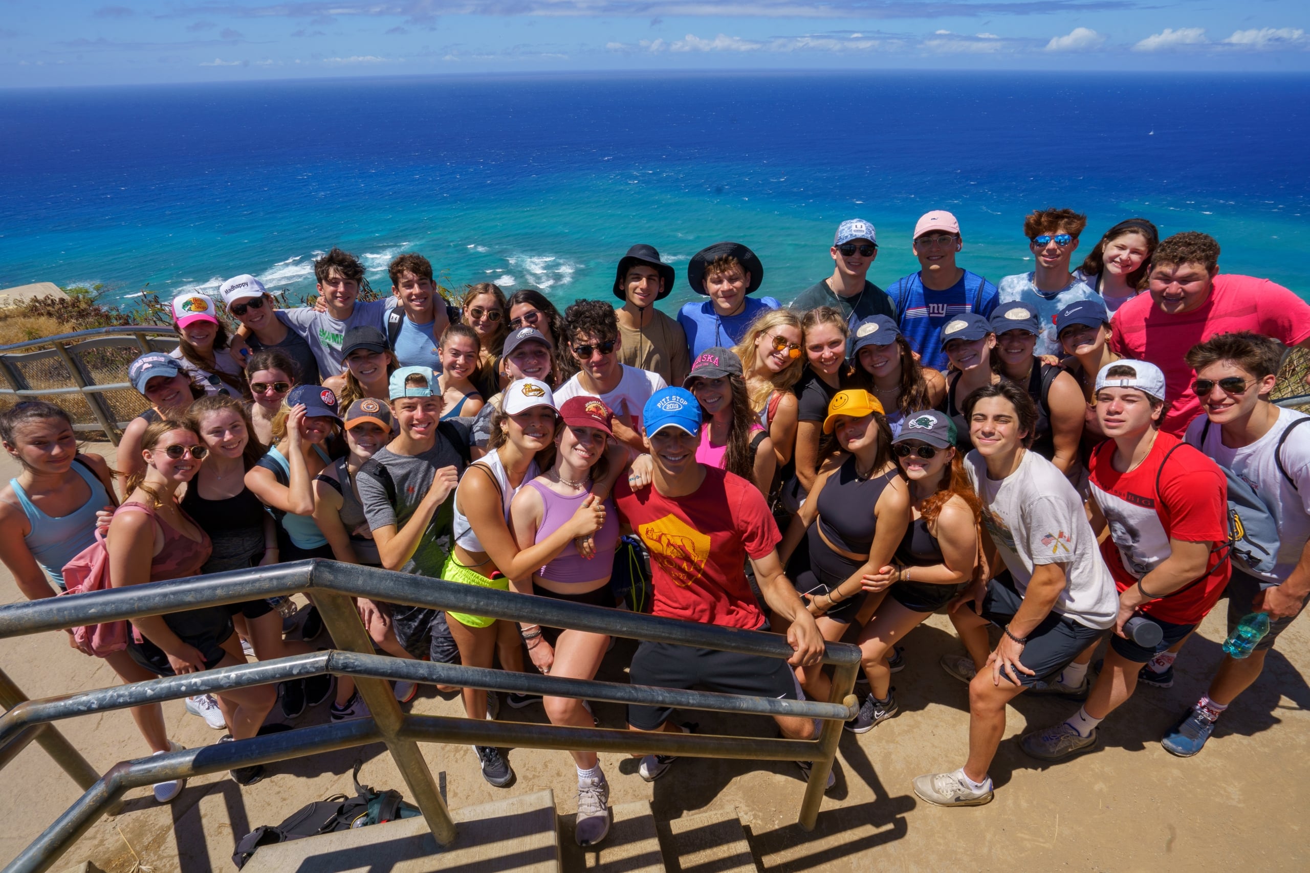 Large group of smiling people posing on a coastal lookout with the ocean in the background.