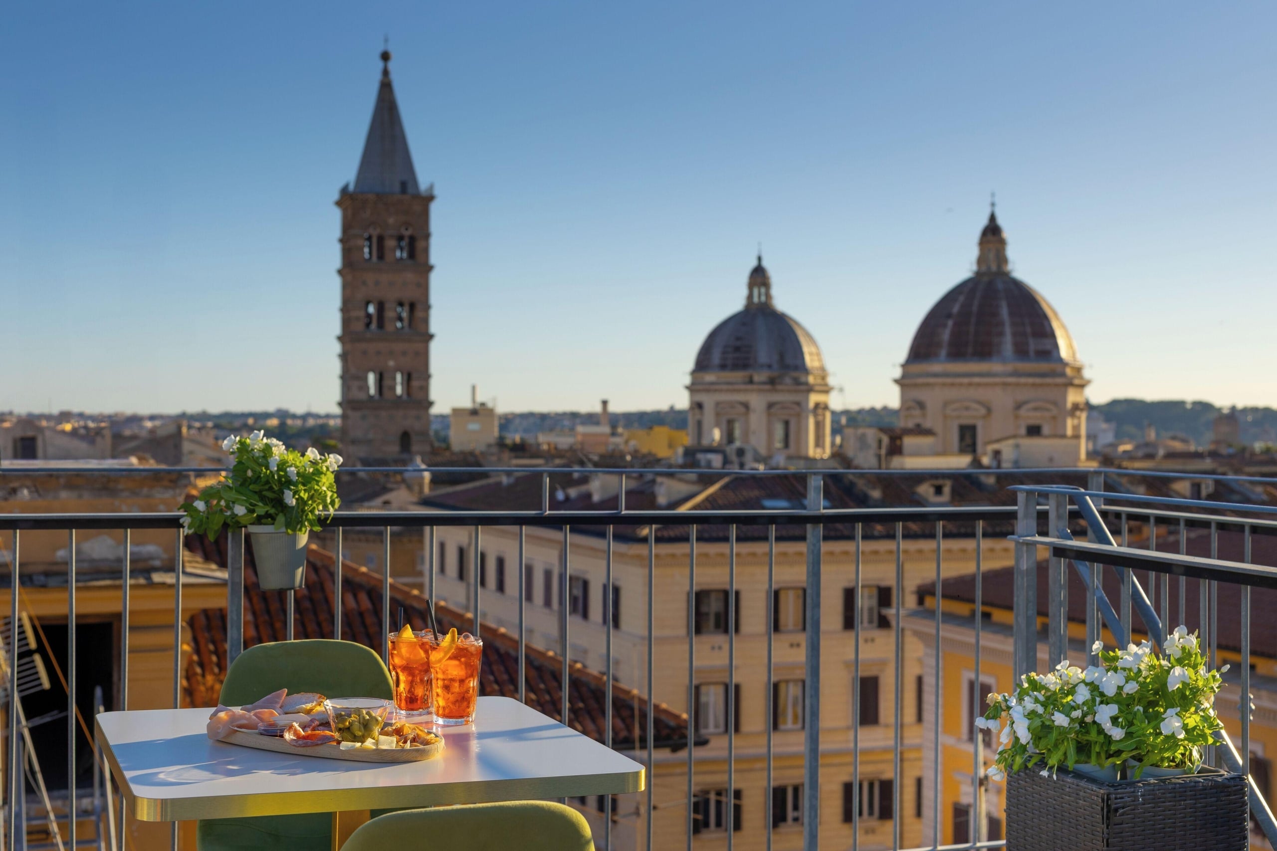 Rooftop terrace table with snacks and orange drinks, green chairs, and potted white flowers, overlooking a city with domed churches and a bell tower at sunset.