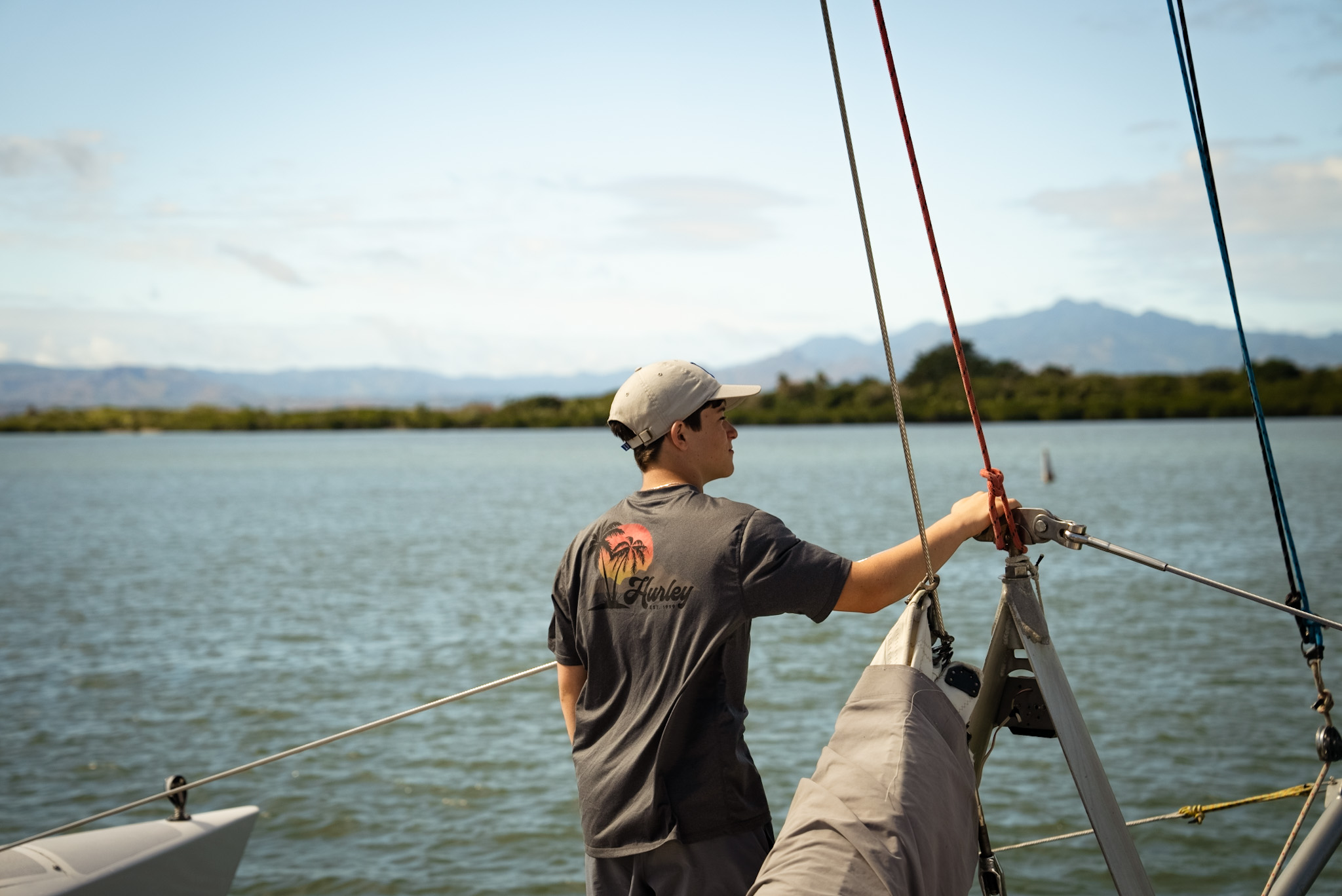 Man standing on a sailboat, holding rigging ropes with a lake and distant mountains in the background.