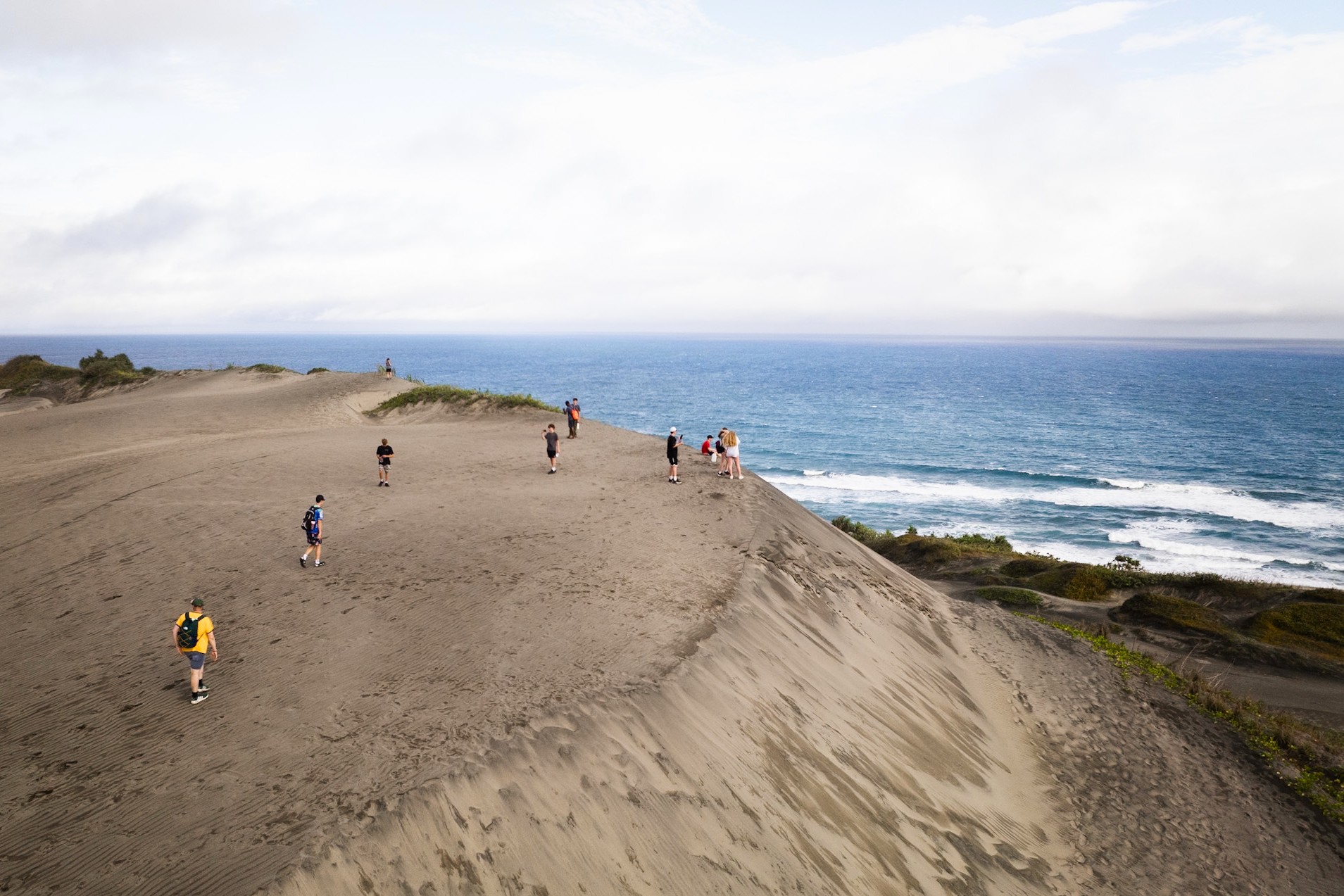 People hike along a wide sandy coastal dune with the sea in the background under a bright sky.