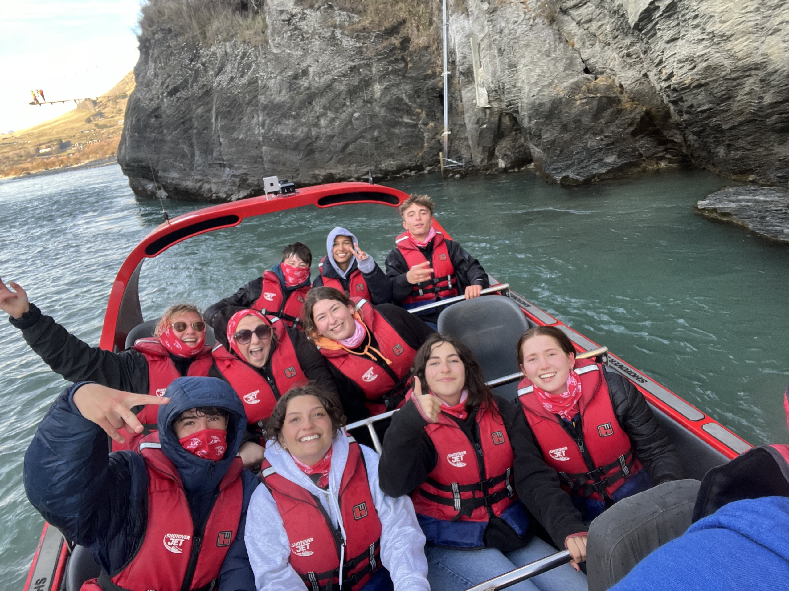 Group of smiling travelers wearing red life vests on a red speedboat, with rocky cliffs and water in the background.