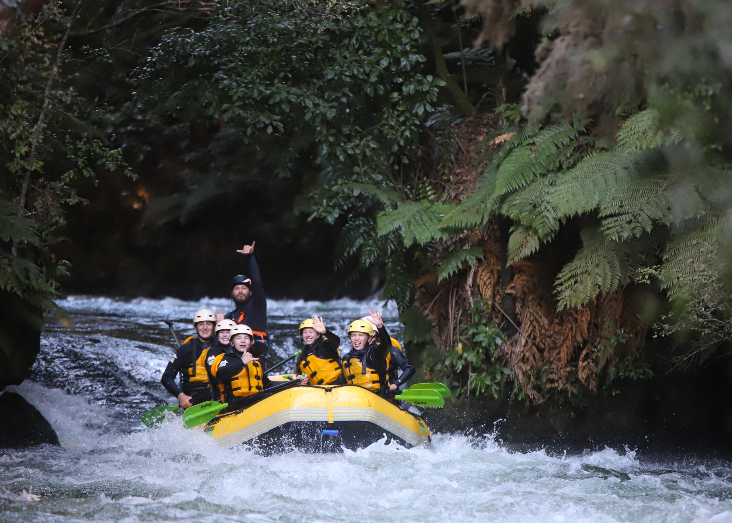 Group of people in yellow life jackets raft down a whitewater river, surrounded by dense green foliage and ferns.