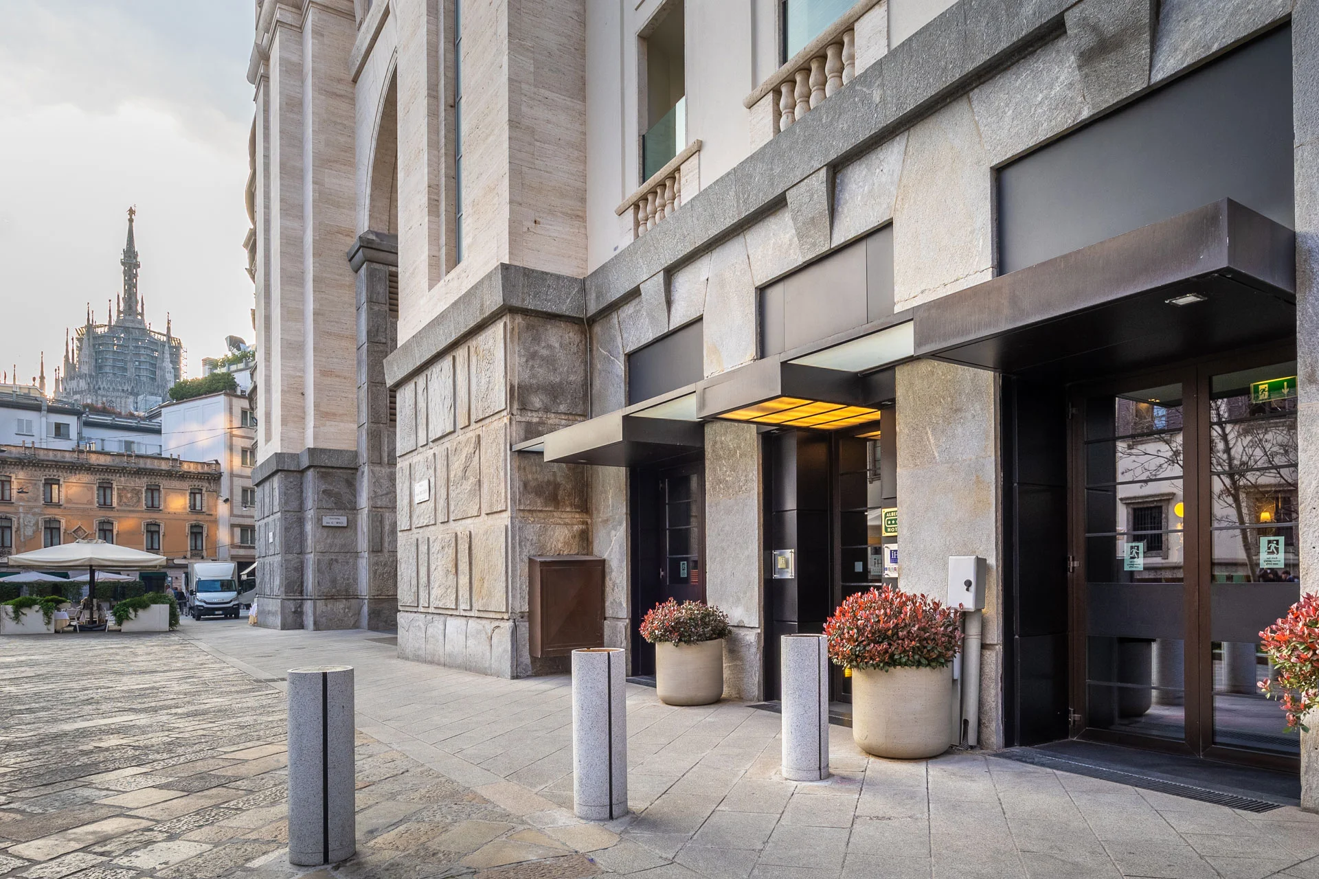 Stone building with glass doors and welcoming yellow-lit entrance, large potted plants, and urban square; cathedral spire visible in distance on the left.