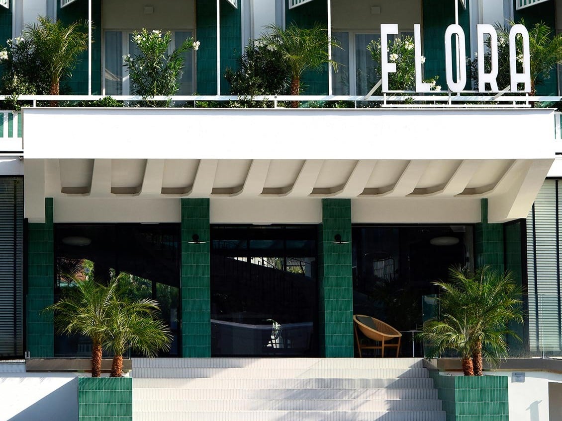 Hotel entrance with a white canopy, green tiled columns, glass doors, and palm trees flanking the stairs with a large 'ELORA' sign above.