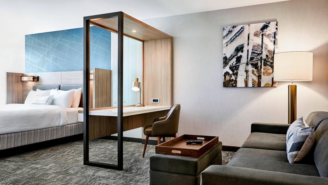 Modern hotel suite: bed to the left, black-framed partition with built‑in desk, chair, and a gray sofa with a wooden tray in the living area.