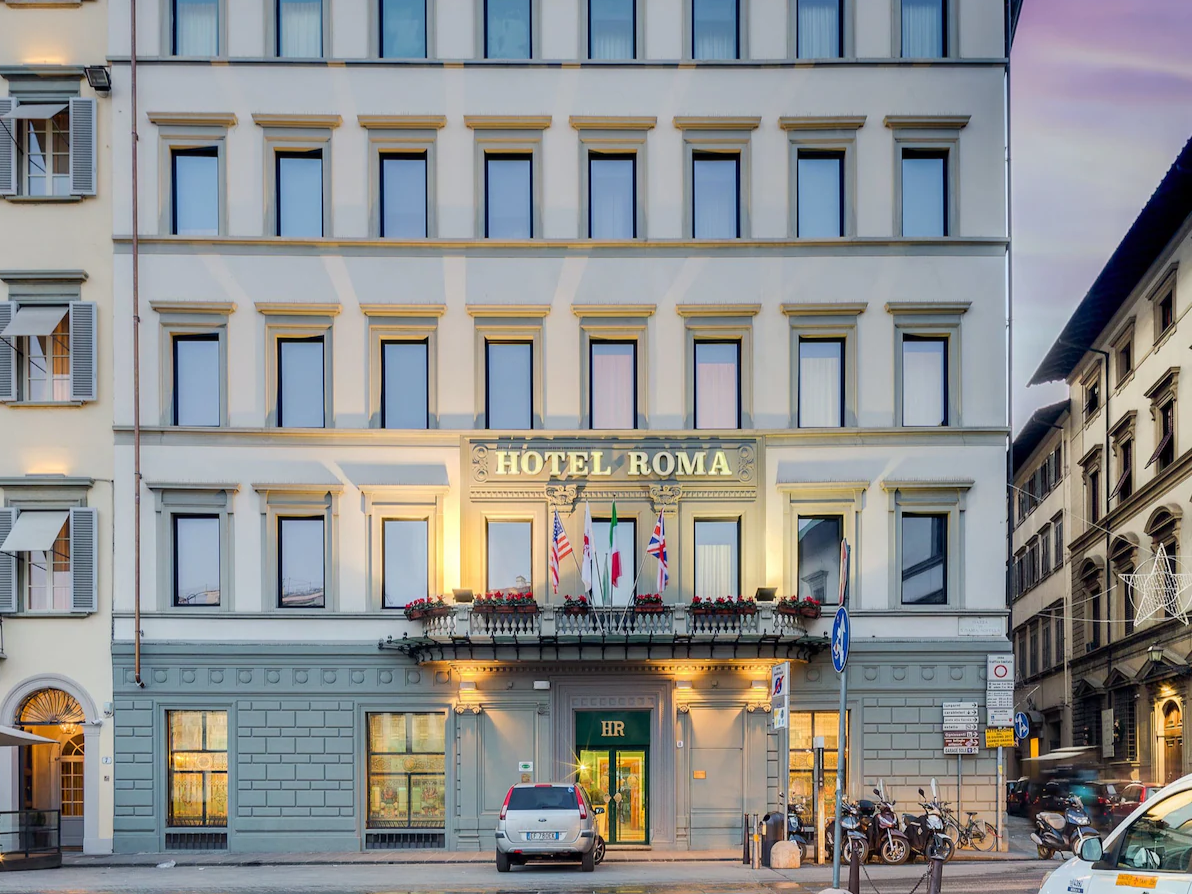 Facade of Hotel Roma with illuminated entrance, flagpoles, and flower boxes on a city street.