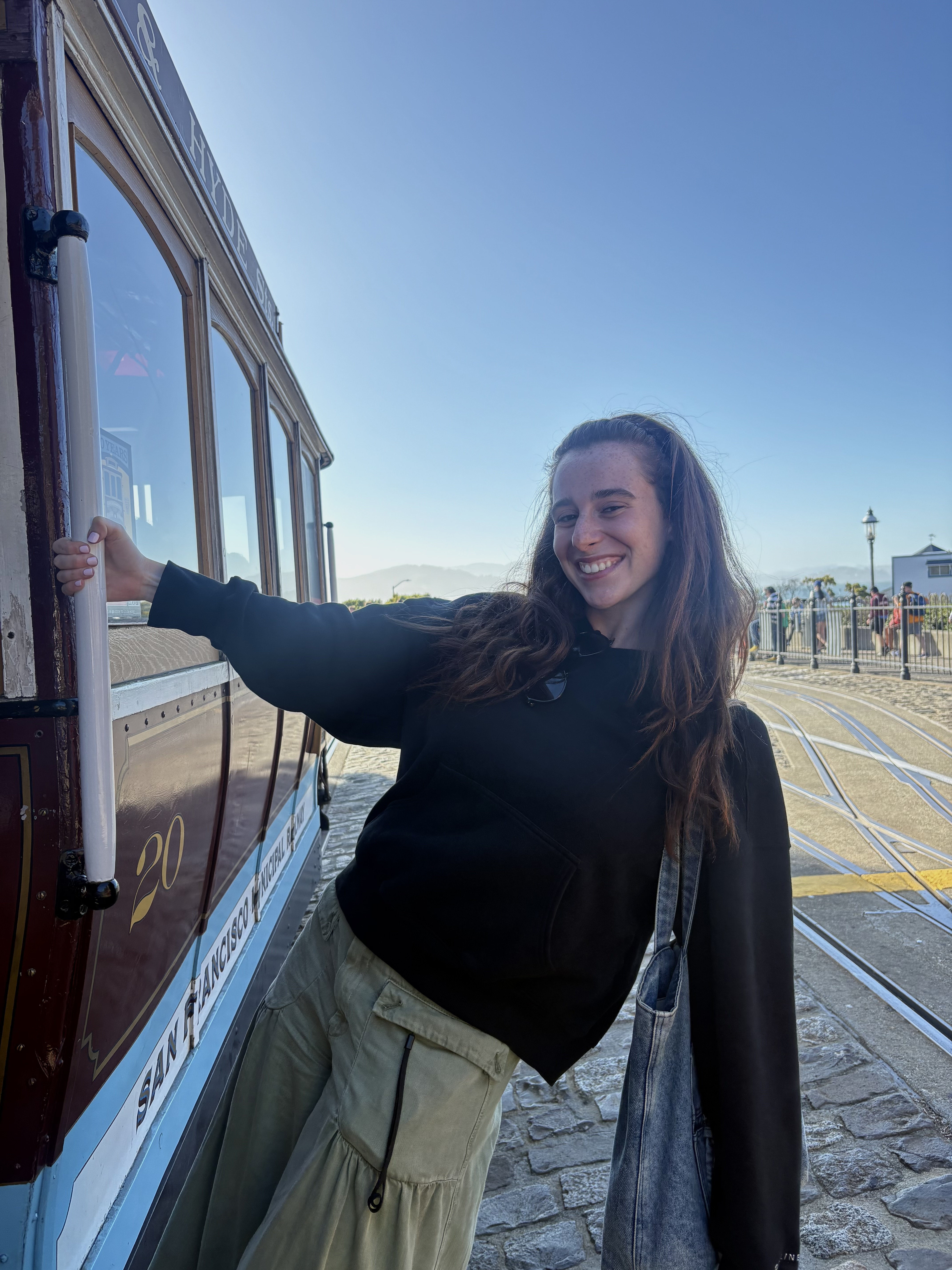 Young woman smiling while posing with a vintage tram on a sunny street, holding a pole with one arm.
