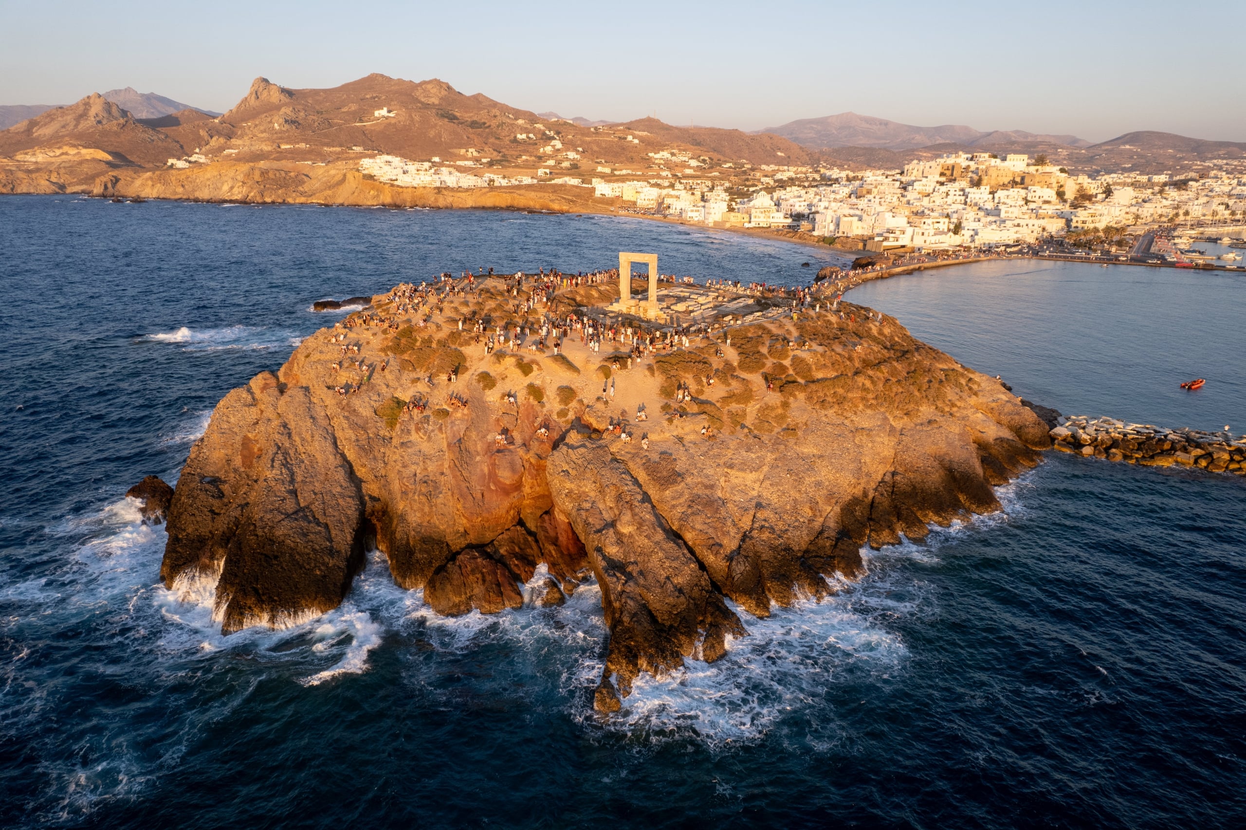 Aerial view of a rocky coastal promontory crowded with people around a rectangular stone arch overlooking the sea and a town beyond.