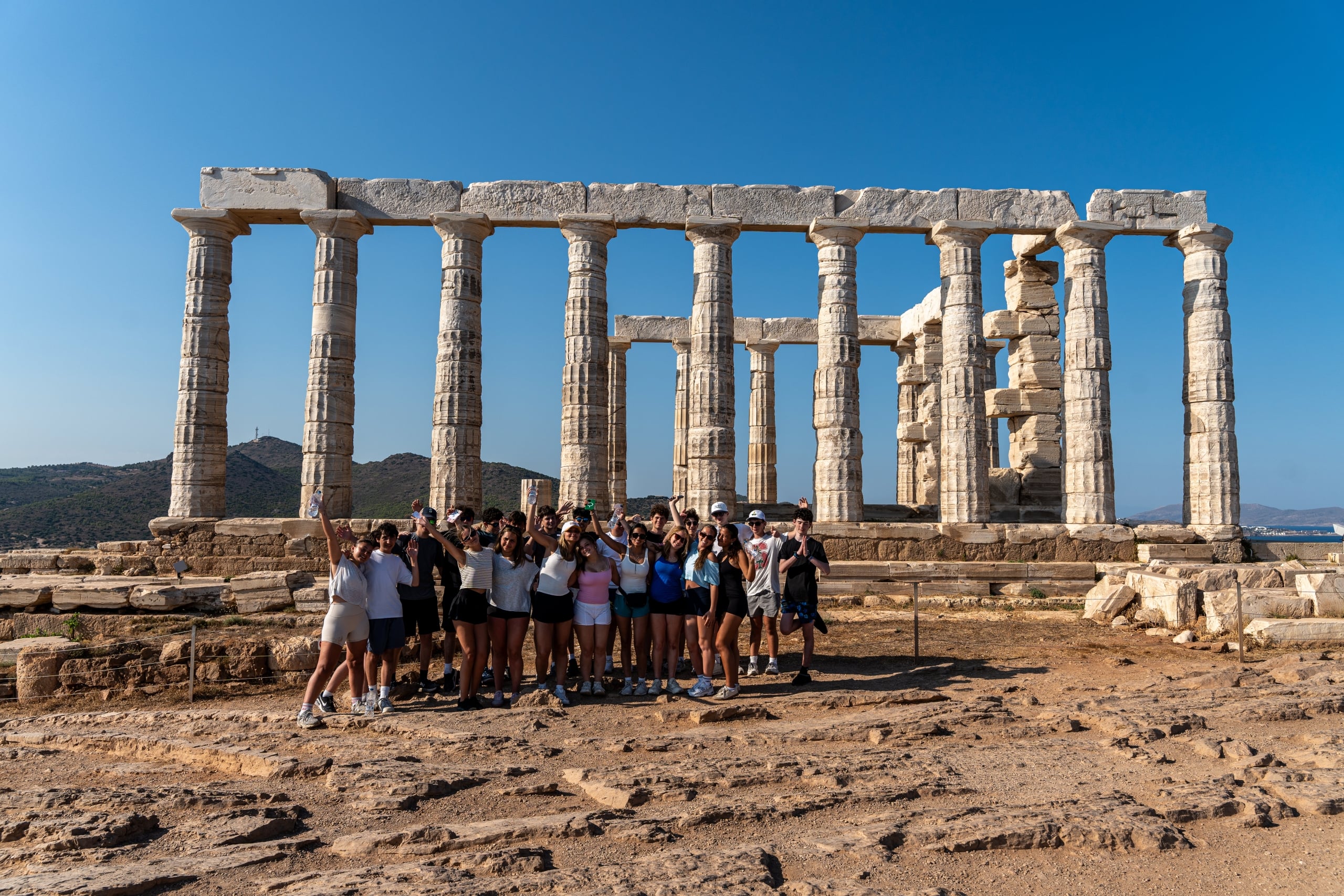 Group of tourists posing in front of ancient stone columns under a clear blue sky, at a ruin site.”