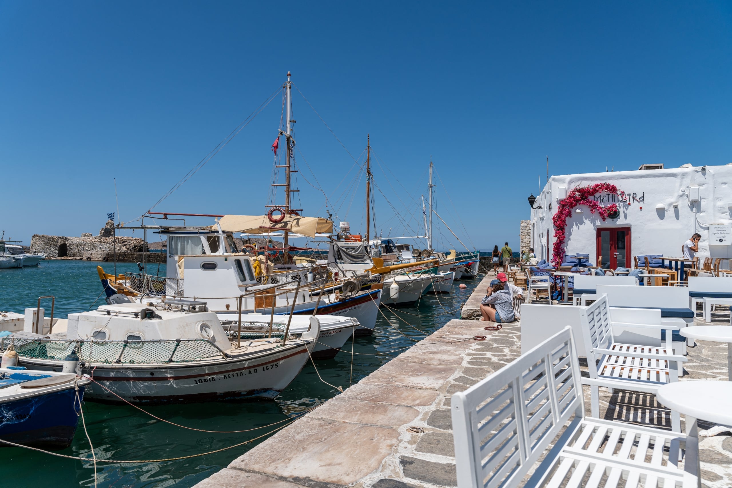 Harbor scene with moored sailboats, a stone pier, and a white building with pink bougainvillea against a clear blue sky.