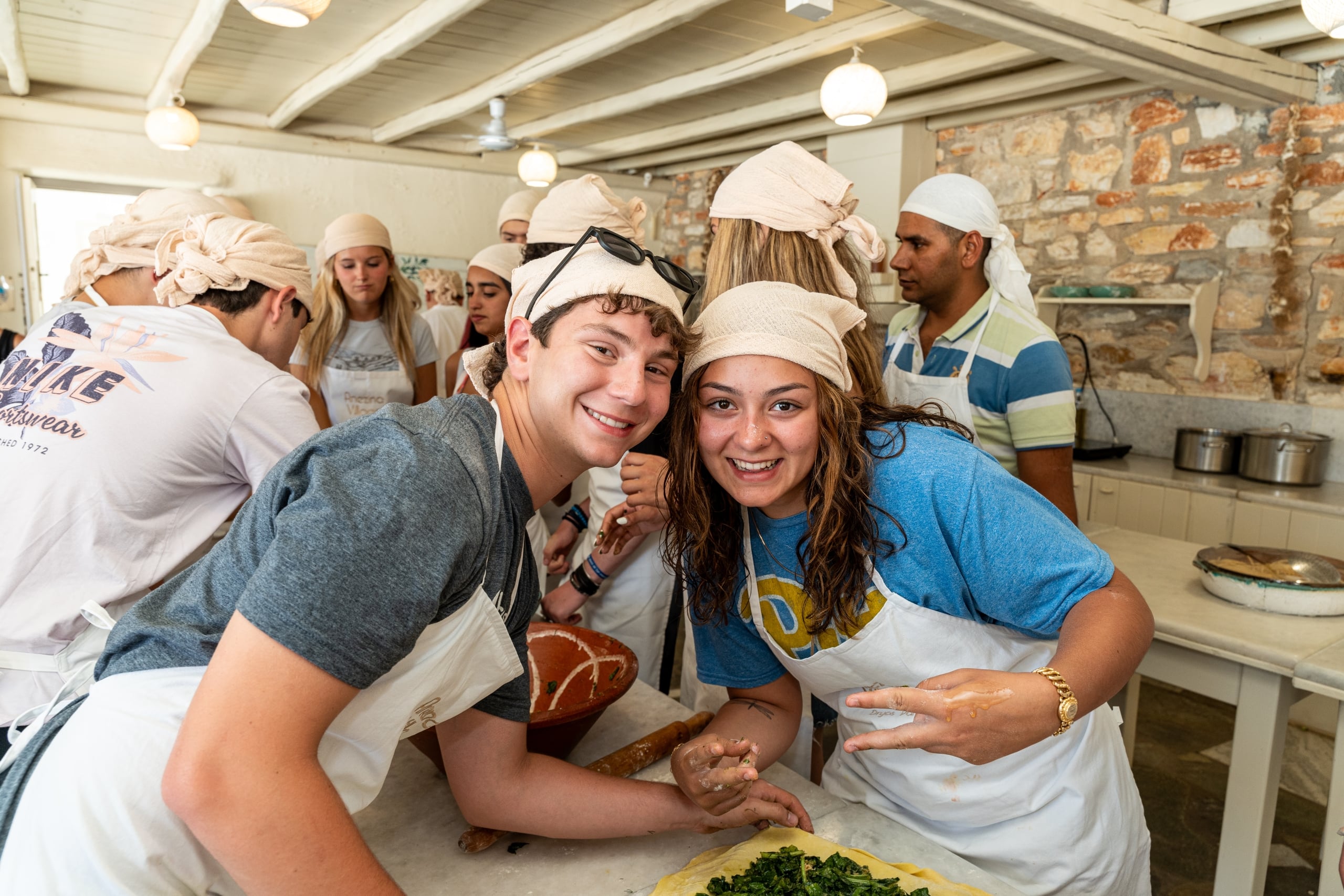 Group of smiling teens in aprons and head coverings gathered around a kitchen prep counter, preparing food together.