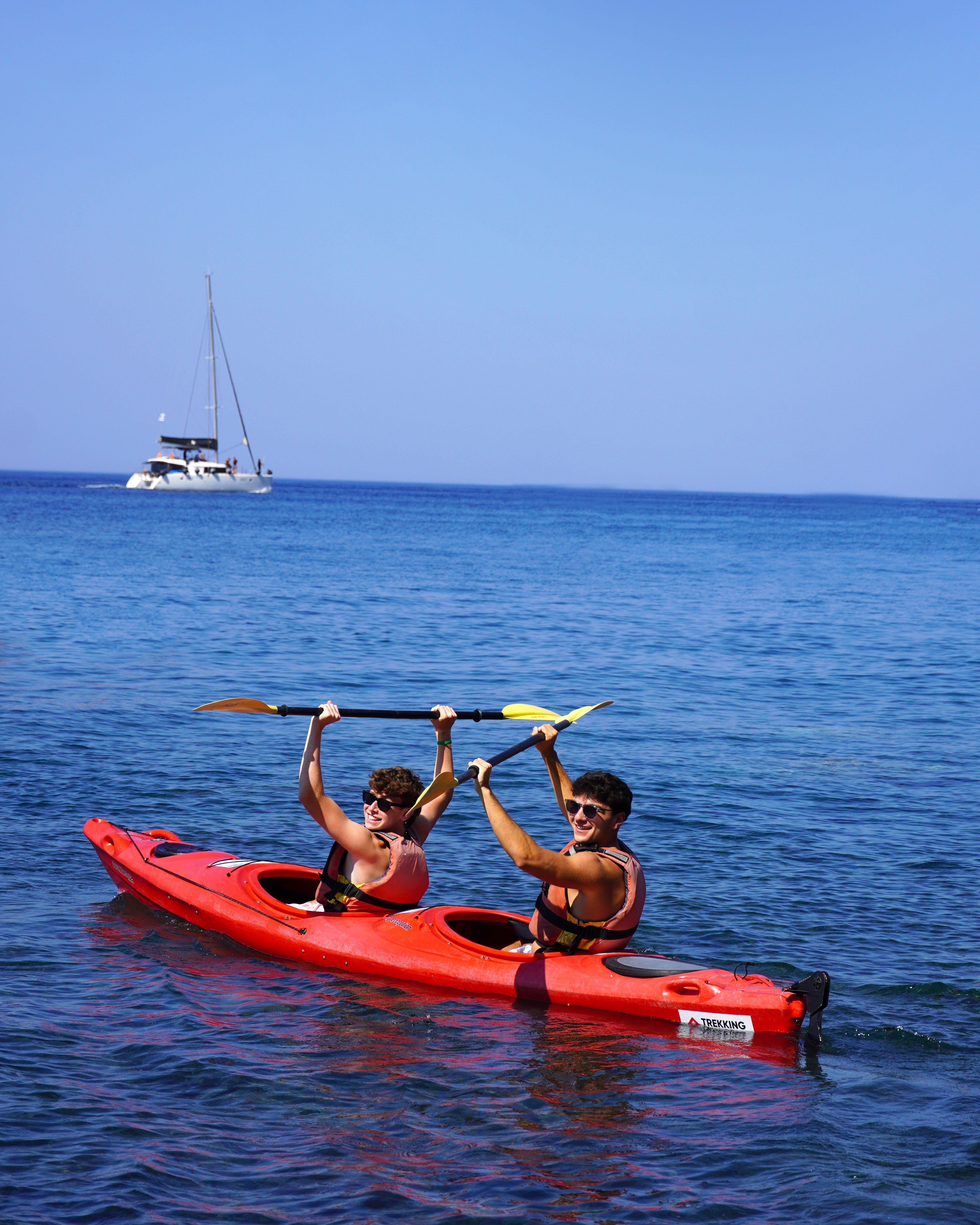 Two people wearing life jackets paddling a red tandem kayak on calm blue water with a distant sailboat in the background.