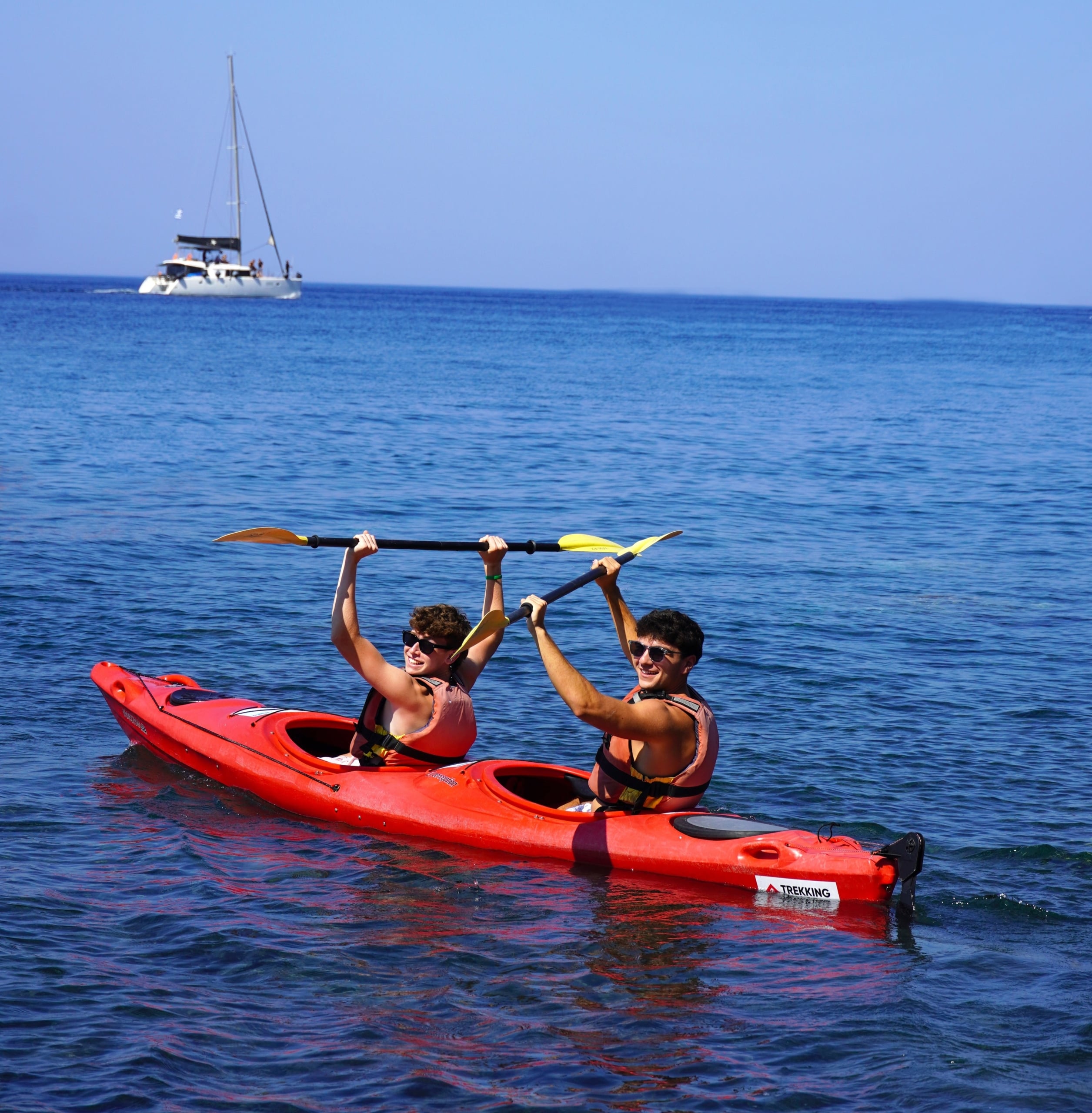 Group of young people in a red tandem kayak raising paddles in the air on calm blue water, with a distant white sailboat on the horizon.