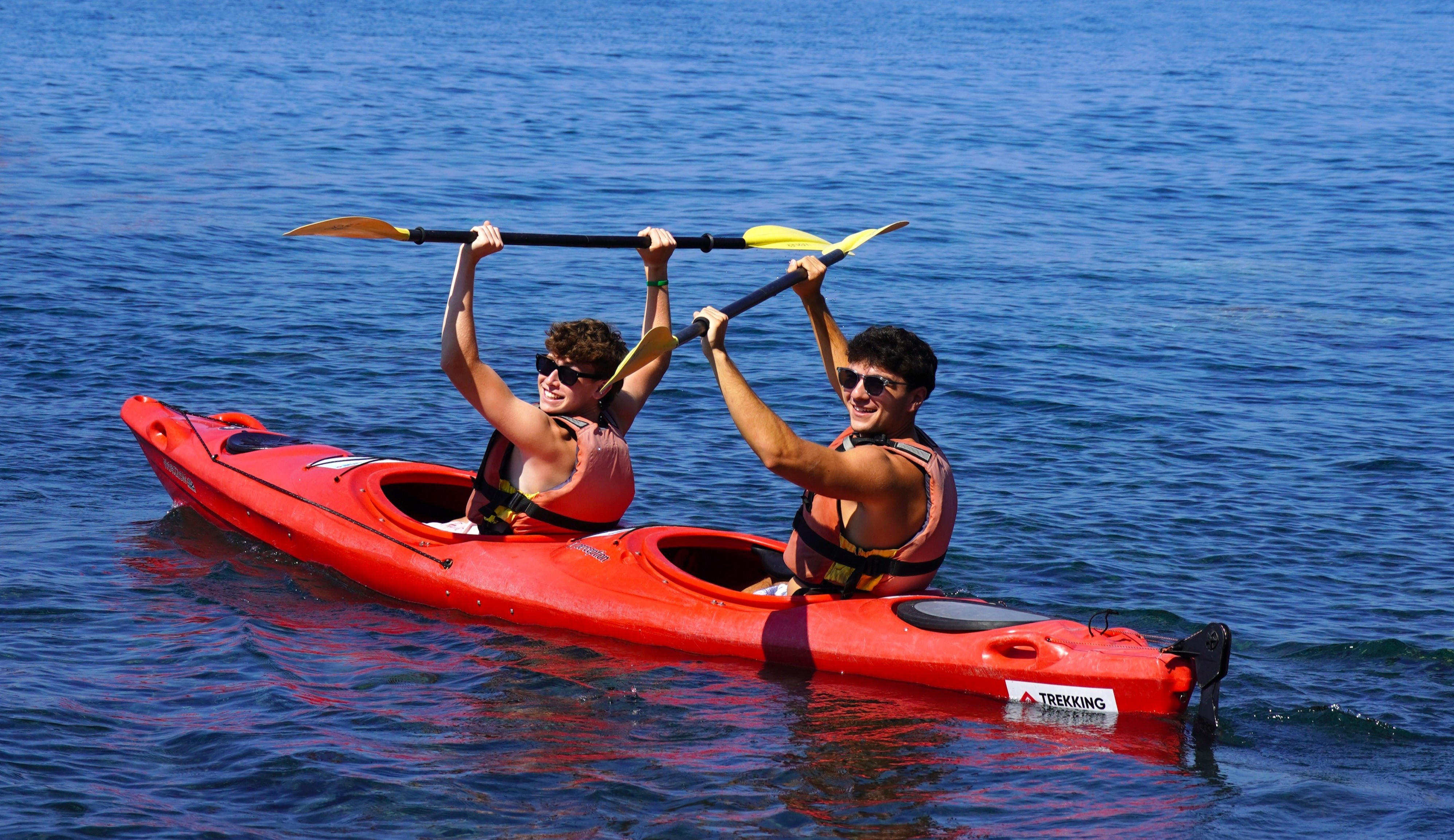 Two people in a red tandem kayak raise their paddles while smiling on blue water.