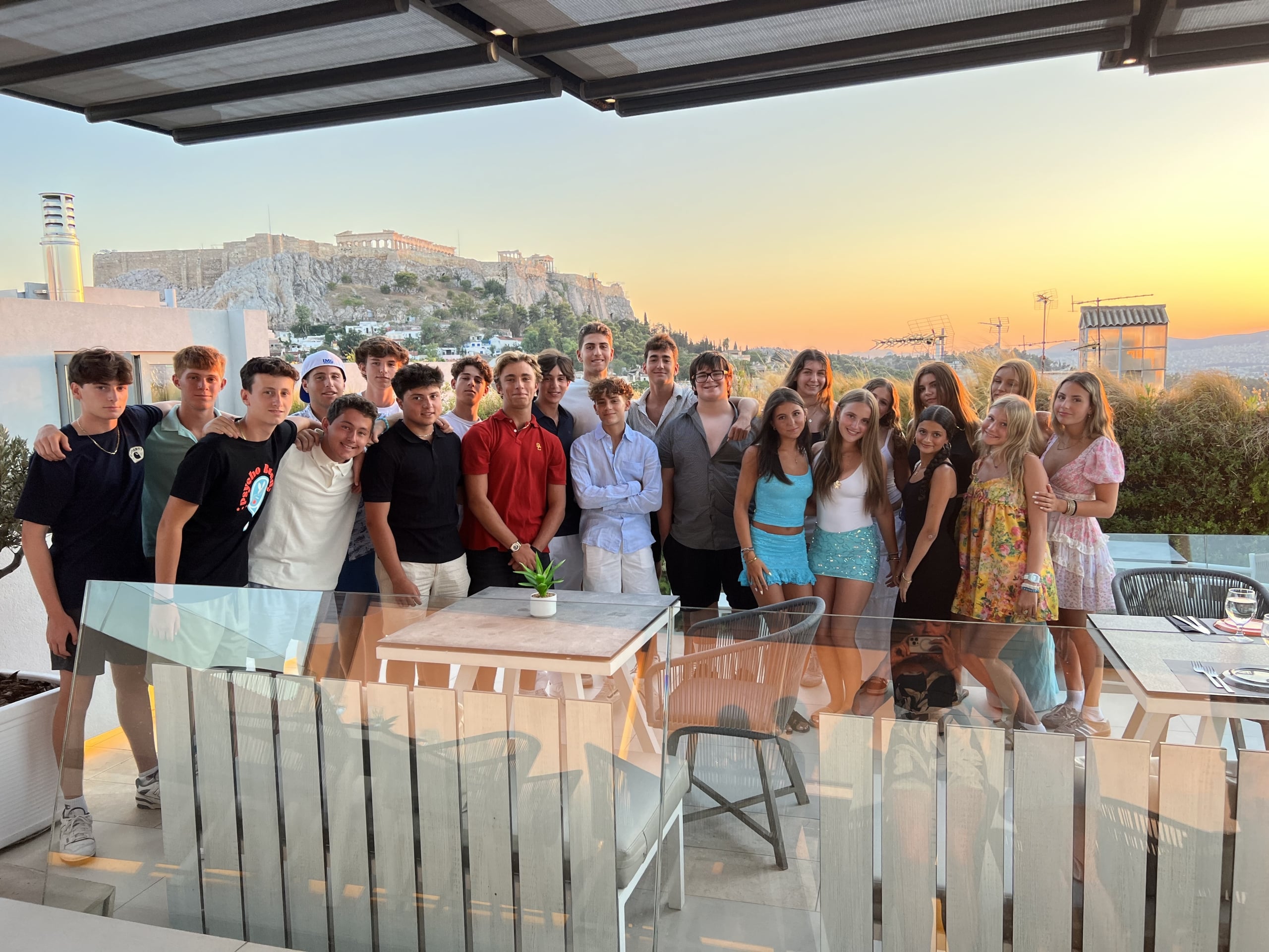 Large group of young people posing together on a sunny rooftop terrace with glass railings, sunset sky, and a hilltop fortress in the background.