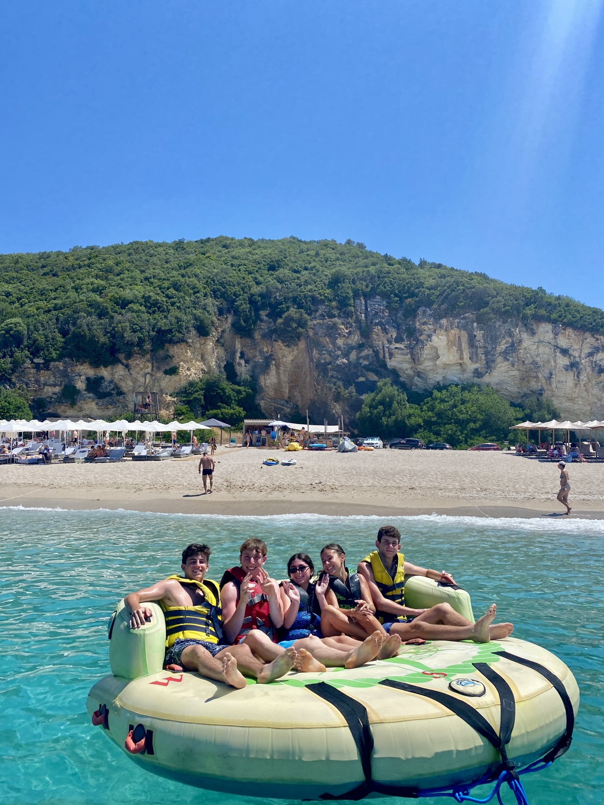 Group of friends wearing life jackets sit on a large inflatable raft in clear turquoise water near a sandy beach with sun umbrellas and a forested hill in the background.
