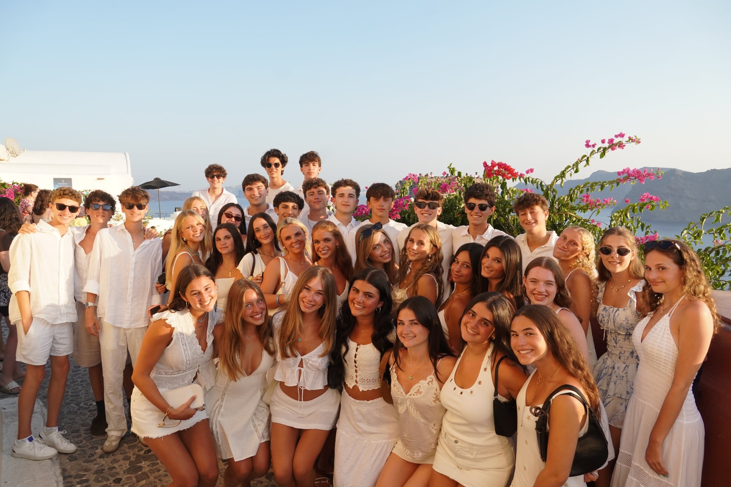 Large group of young people dressed in white posing for a photo on a sunny outdoor terrace with blue sky and flowering bushes in the background.