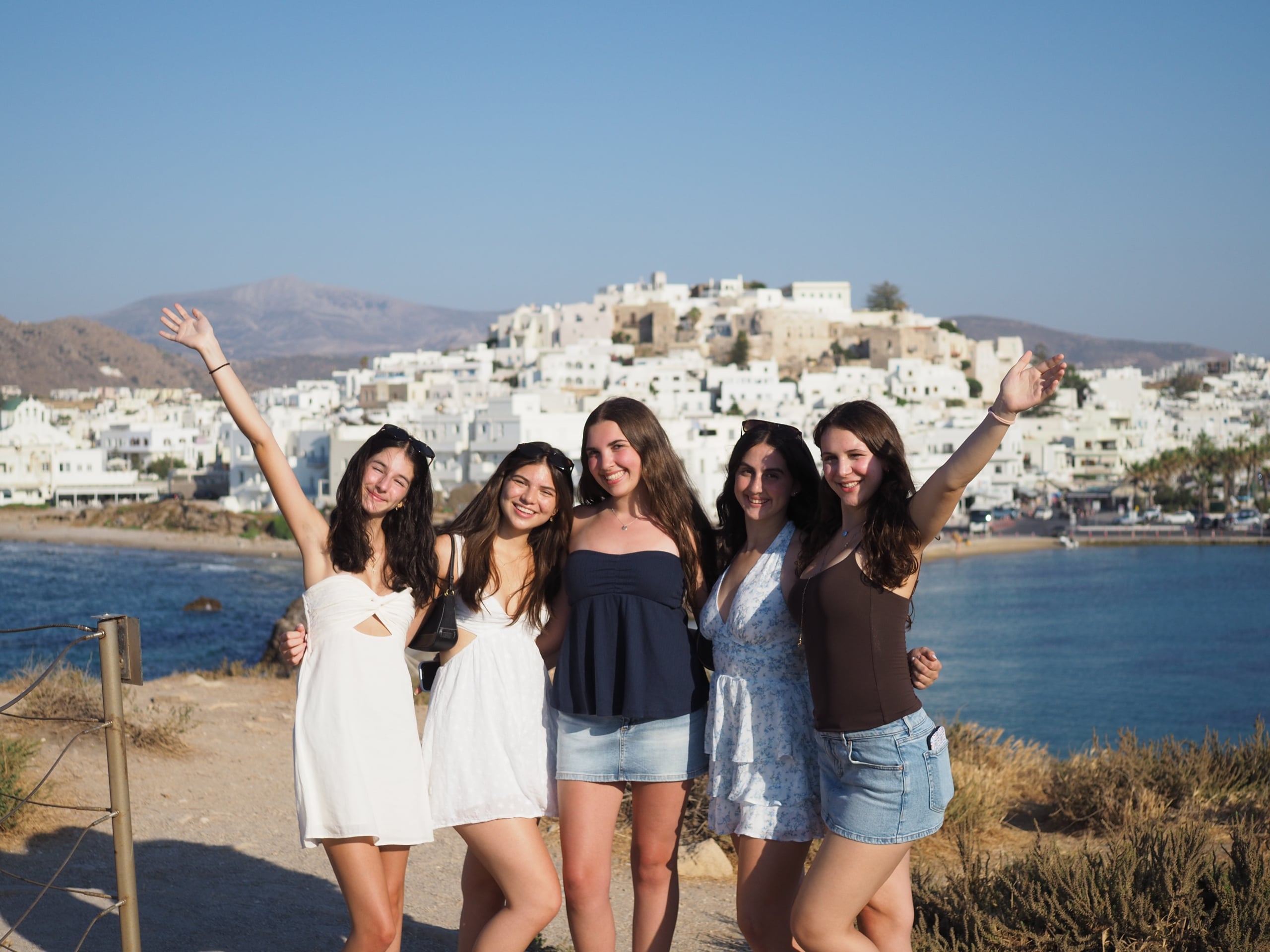 Group of six young women posing with arms raised on a sunny beach, with white hillside town in the background.