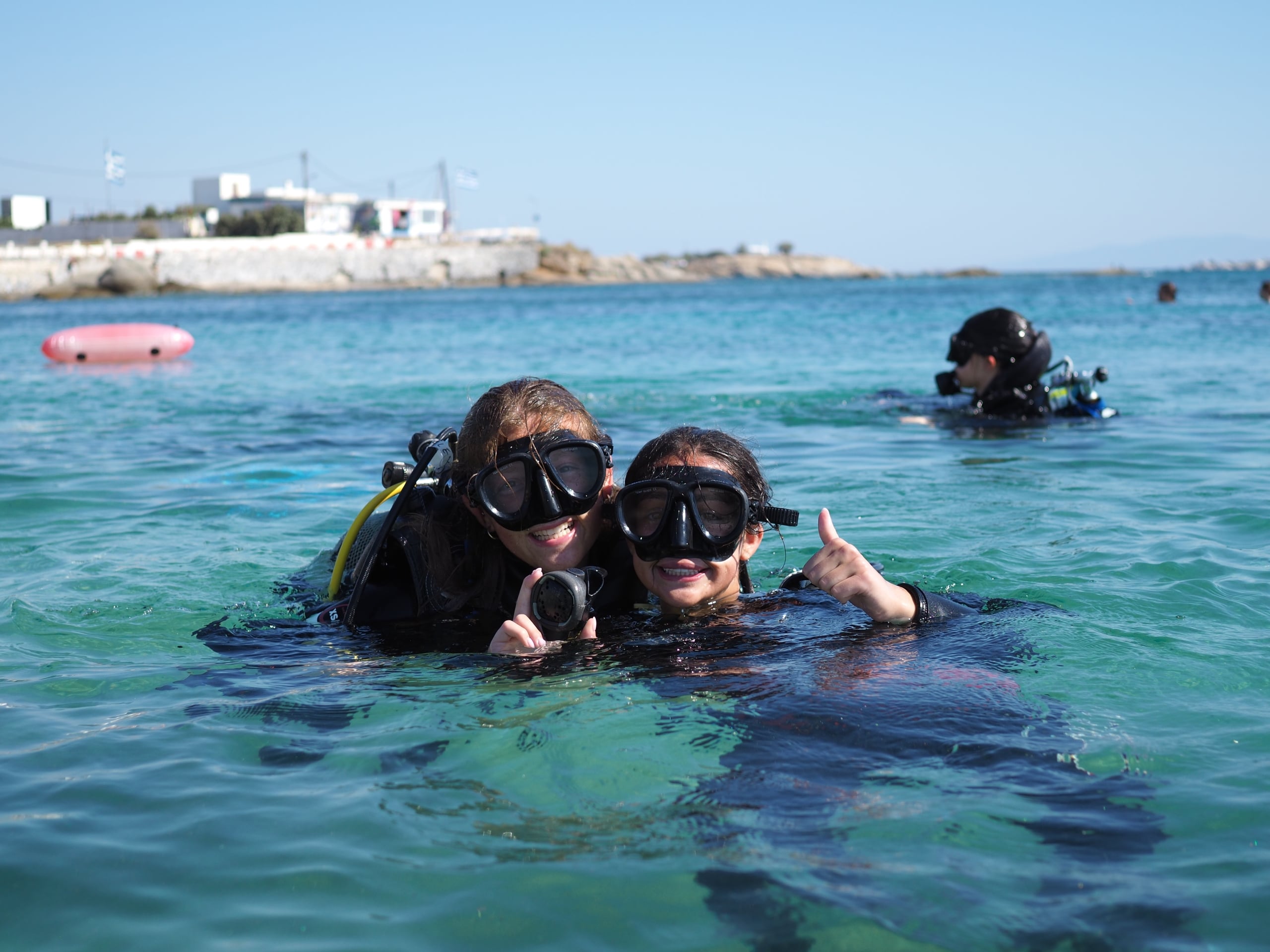 Two snorkelers in the foreground give thumbs up while floating in clear turquoise water, with a rocky shoreline and buildings in the background.