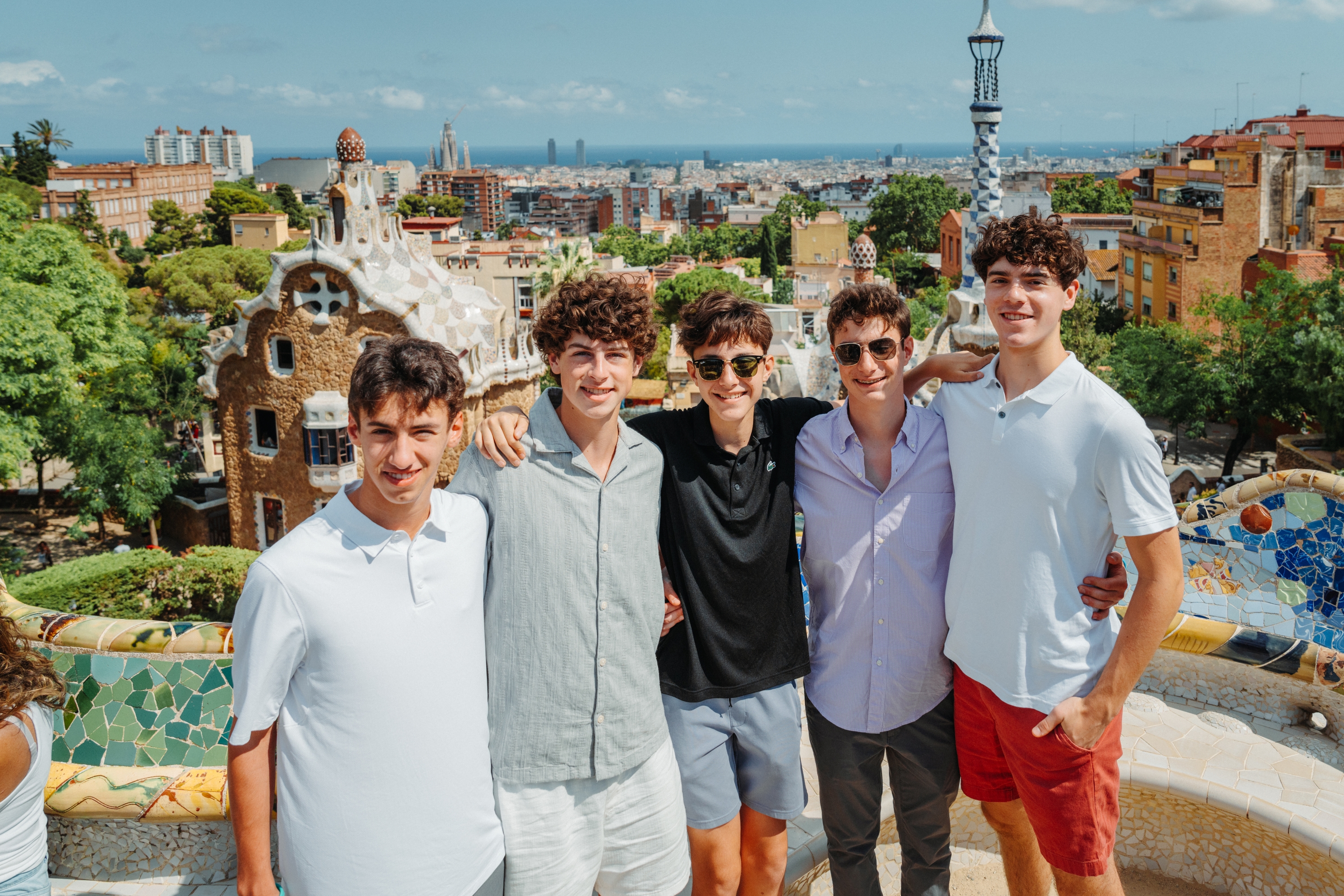 Five young men posing together on a Gaudí mosaic bench at Park Güell, Barcelona, with the city skyline and sea in the background.