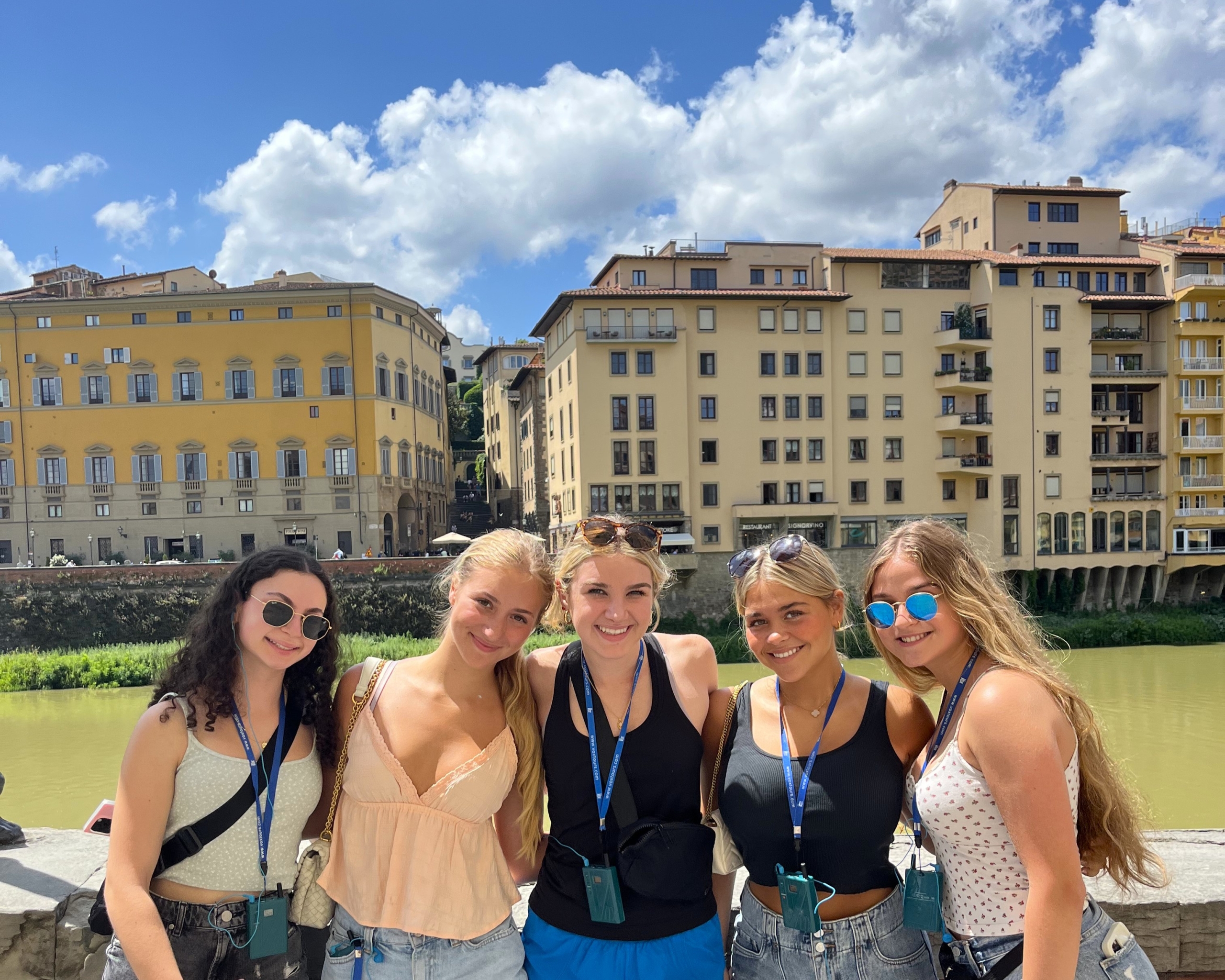 Group of five young women smiling for a selfie in front of a river and yellow apartment buildings on a sunny day.