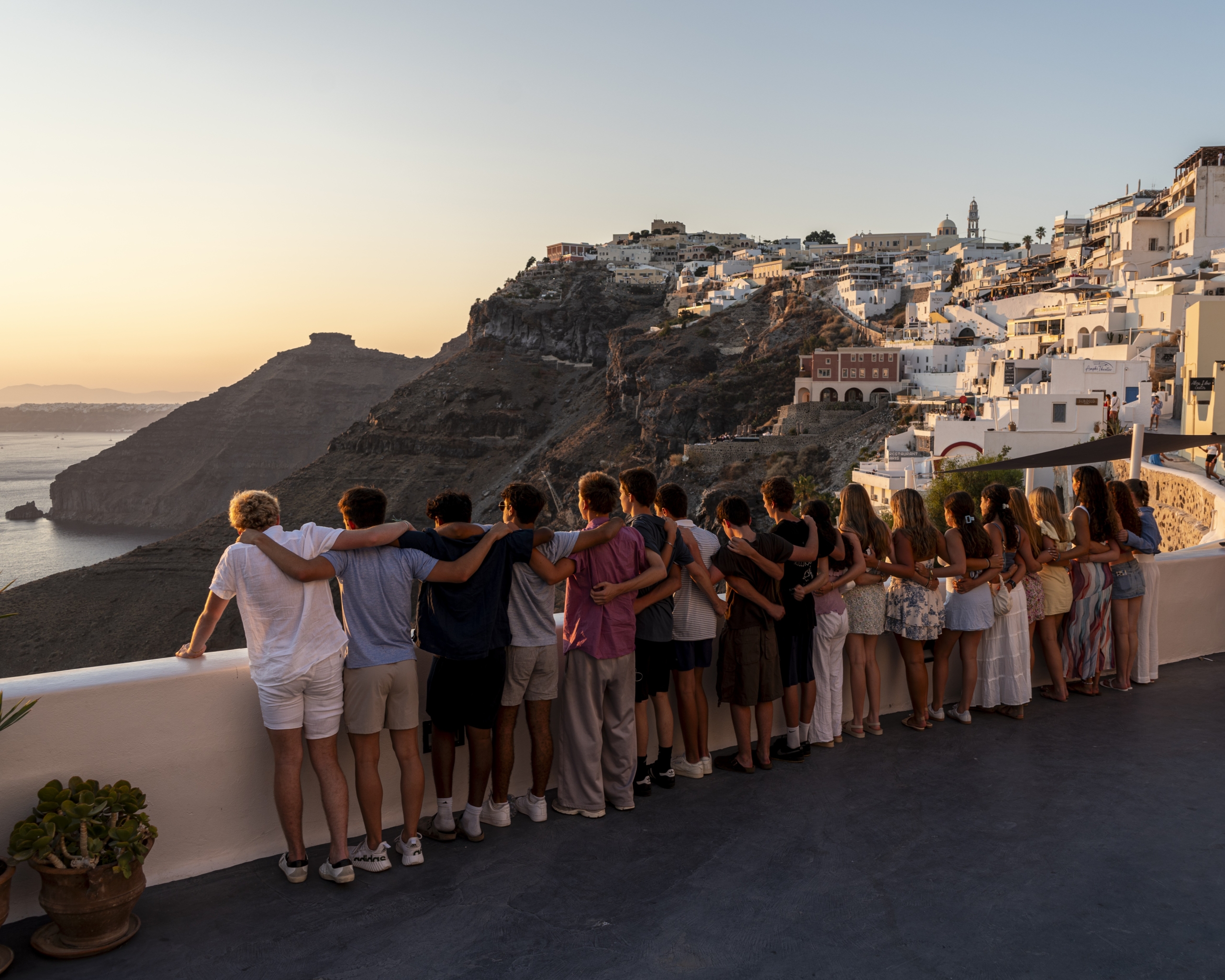 Group of people with arms around each other along a railing, watching a cliffside town at sunset with white-washed buildings on the slope behind them.