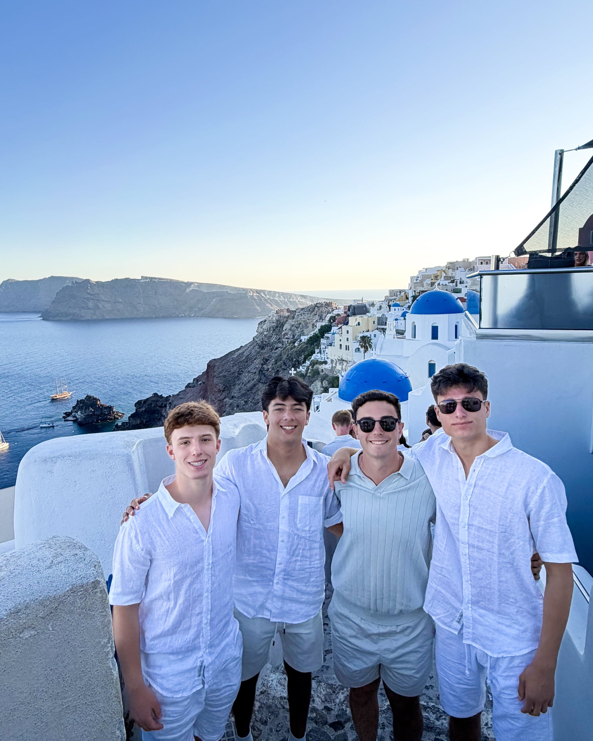 Six young men in white shirts pose together on a cliffside in Santorini, with white buildings, blue domes, and the sea in the background.