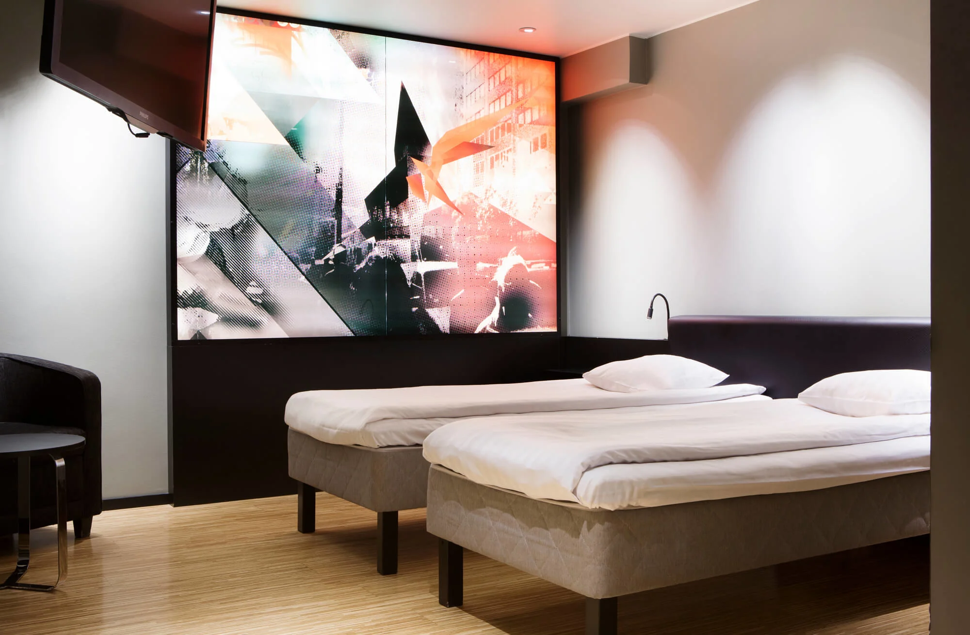 Two twin beds in a modern hotel room with a large abstract mural and a wall-mounted flat-screen TV above.