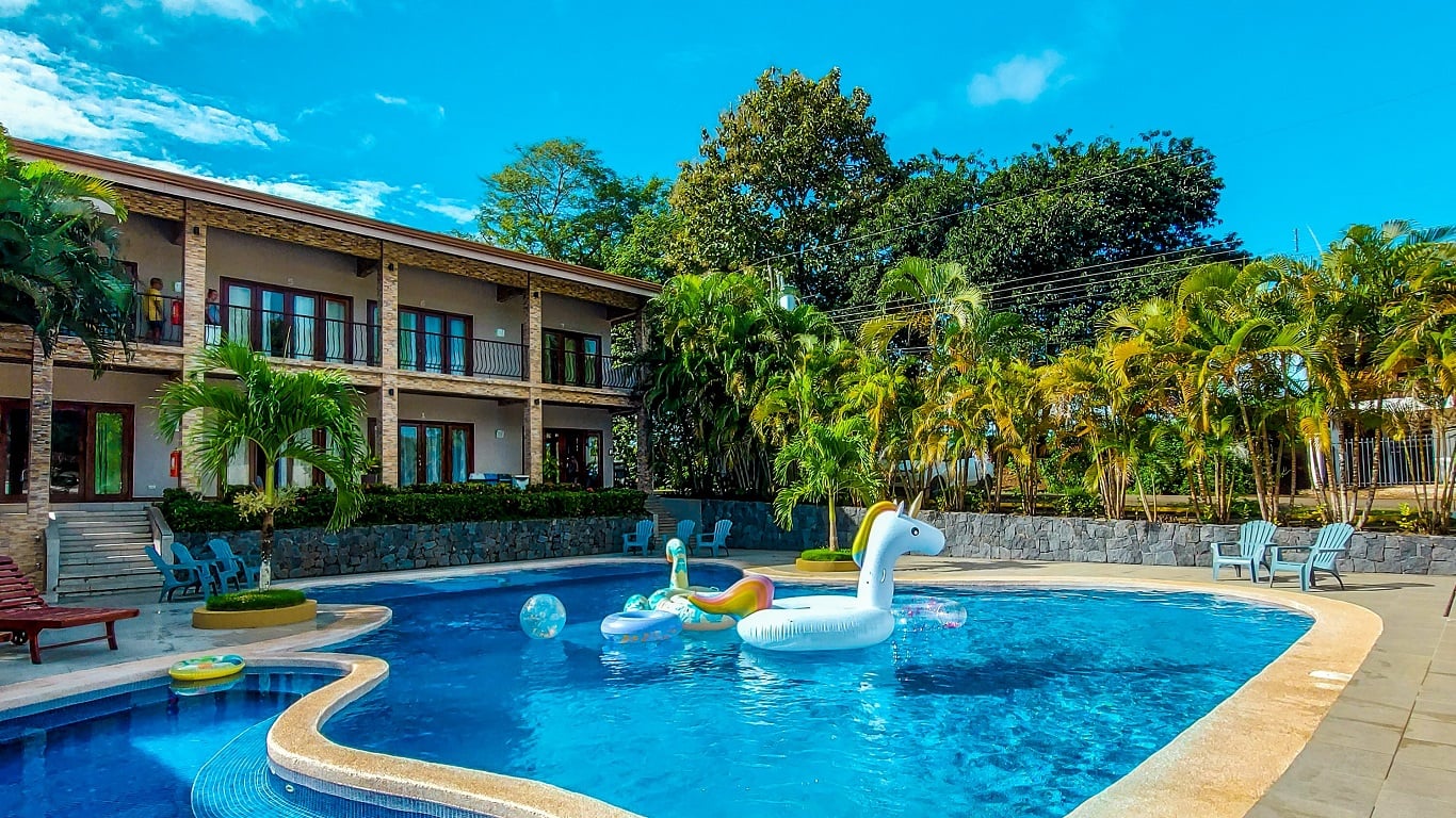 Outdoor hotel pool with inflatable unicorn and colorful floats, palm trees, and a two-story building in the background.