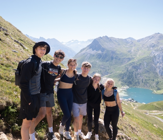 Six friends posing together on a rocky mountain trail with snowy peaks and a turquoise lake in the background on a sunny day.