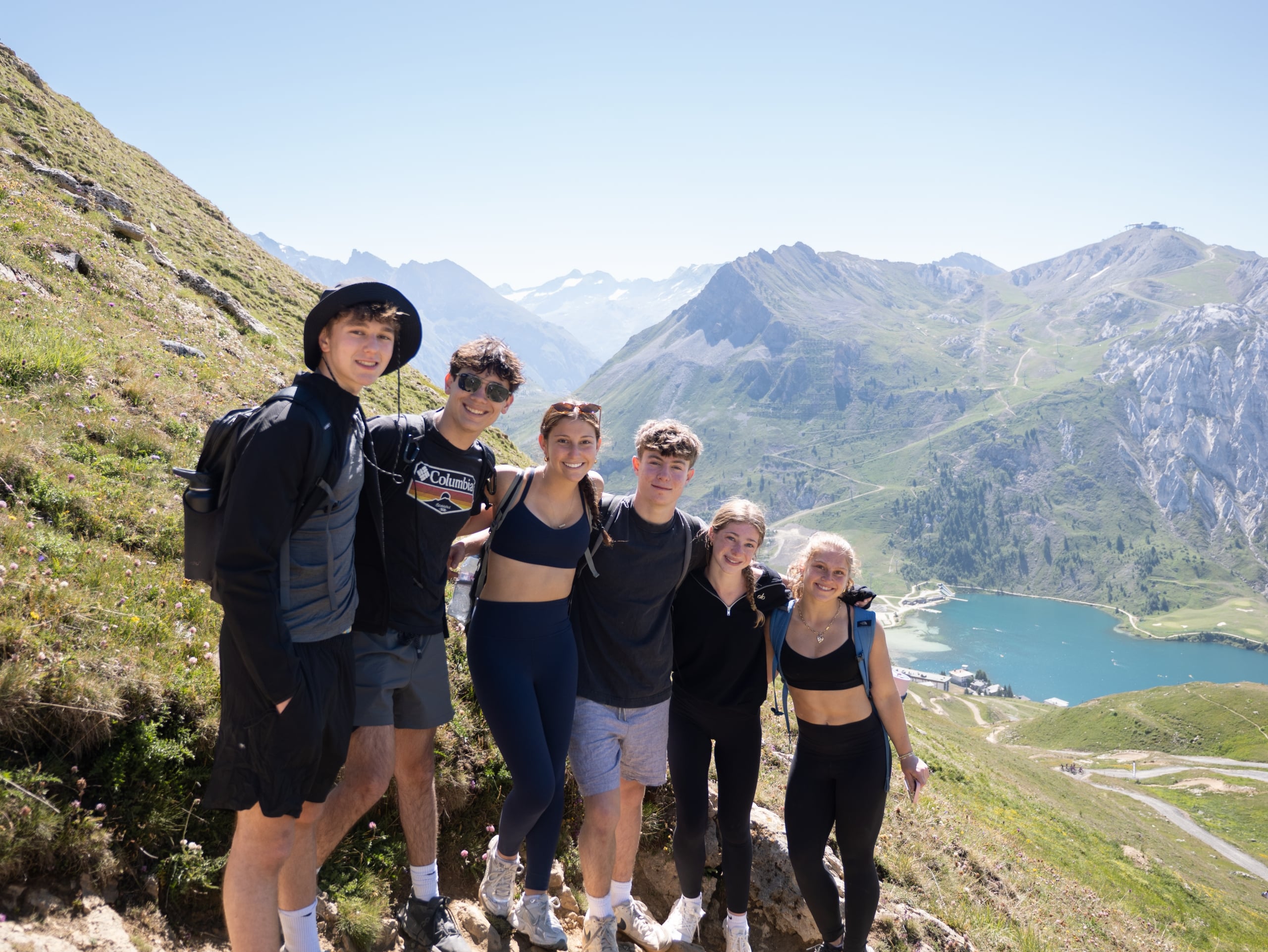 Six friends posing together on a rocky mountain trail with snowy peaks and a turquoise lake in the background on a sunny day.