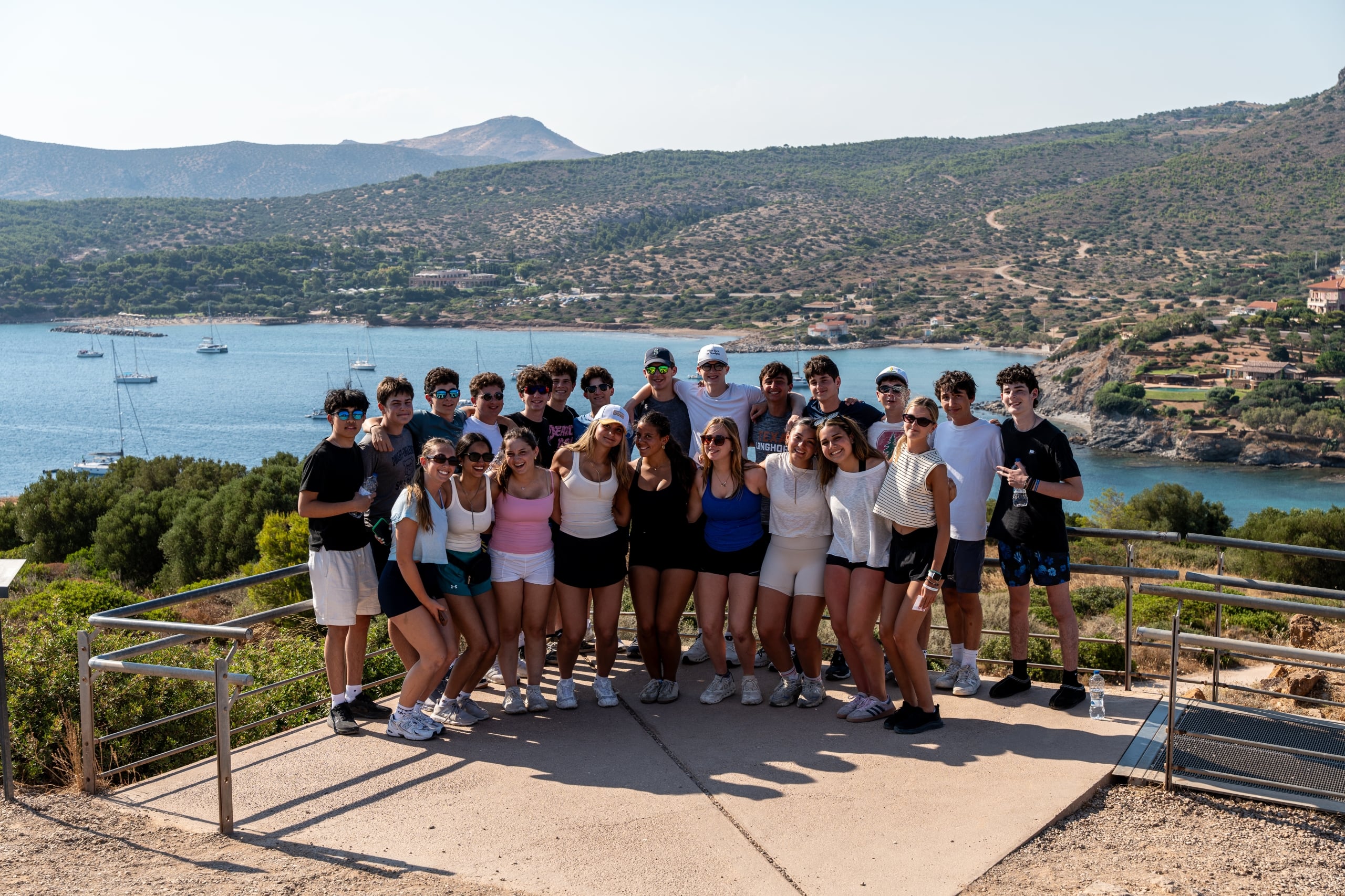 Group of smiling youths posing together on a overlook railing with a coastal bay and hills in the background, boats in the water.