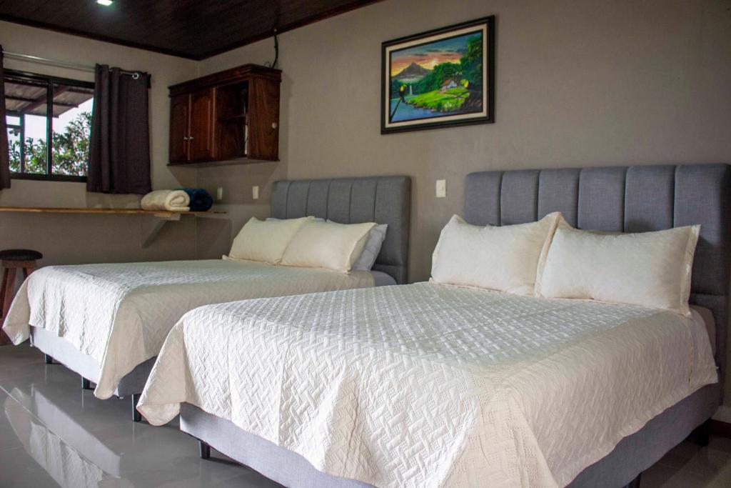 Two queen beds with gray headboards and white quilted bedspreads in a bright hotel room, plus a wall painting above the beds.