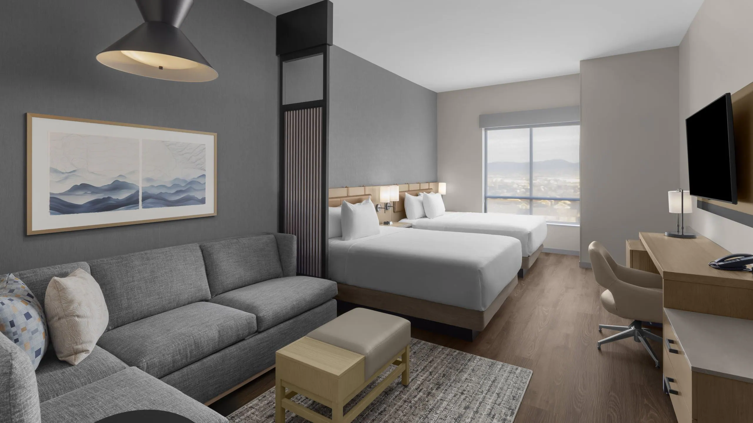 Modern hotel suite: living area with gray sofa, wall art, a divider, two white beds, and a desk near a large window with city view.