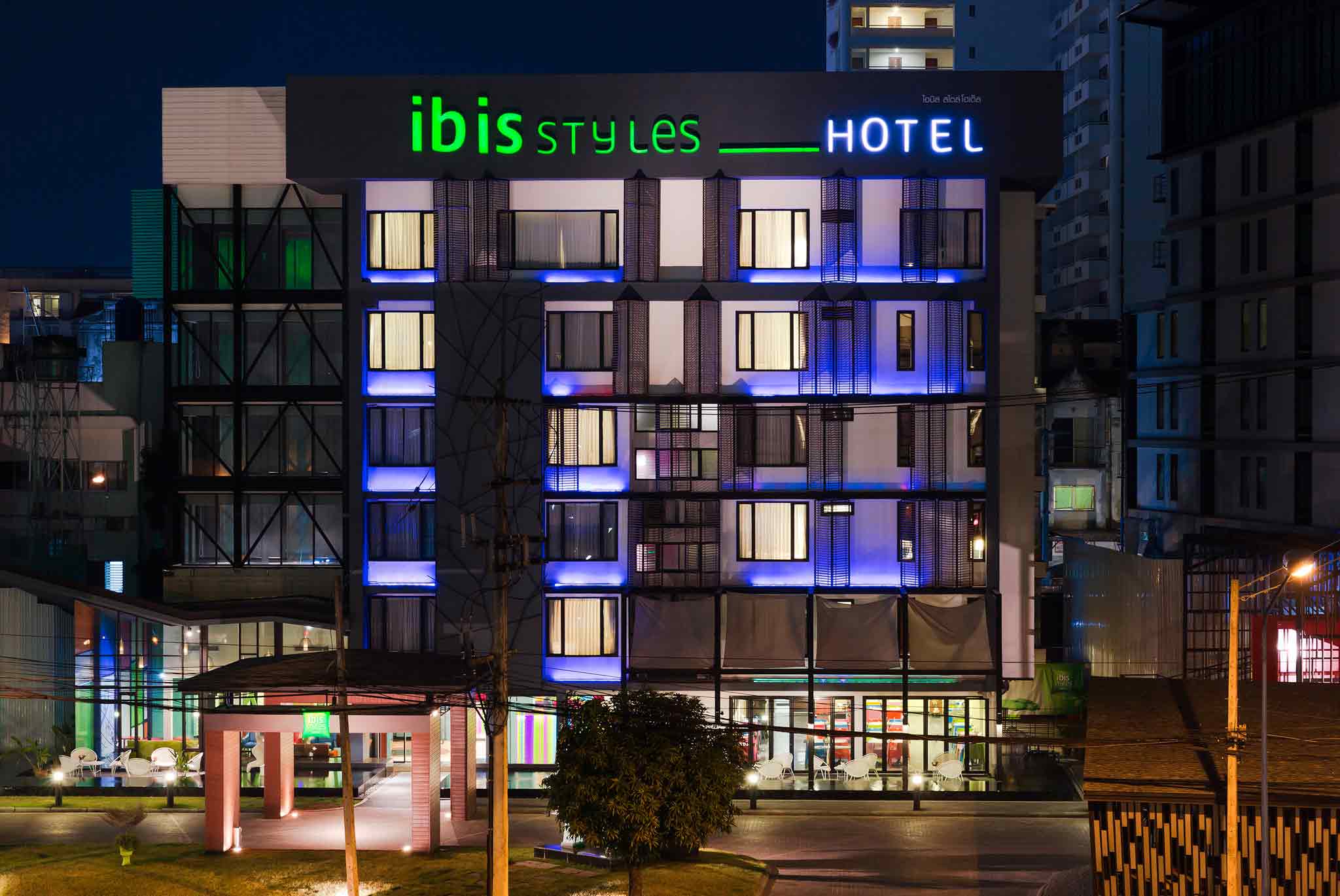Night-time facade of a modern ibis Styles hotel with green 'ibis' and blue 'HOTEL' neon signage and lit windows across floors.