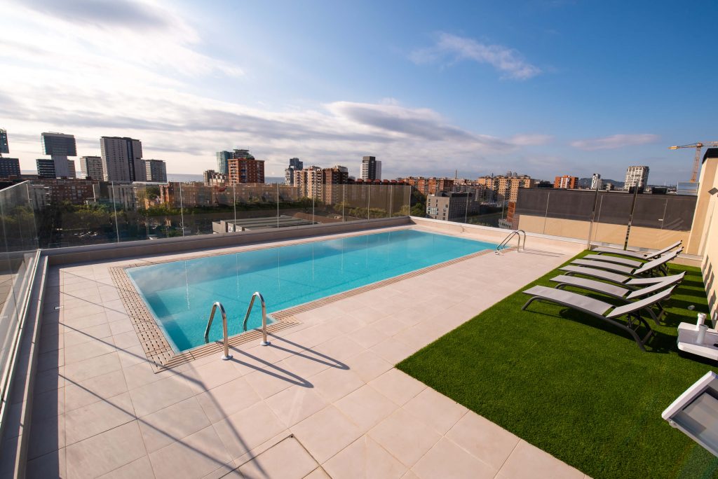 Rooftop pool with a glass railing, ladder entry, and lounge chairs on artificial grass overlooking the city skyline at sunset.