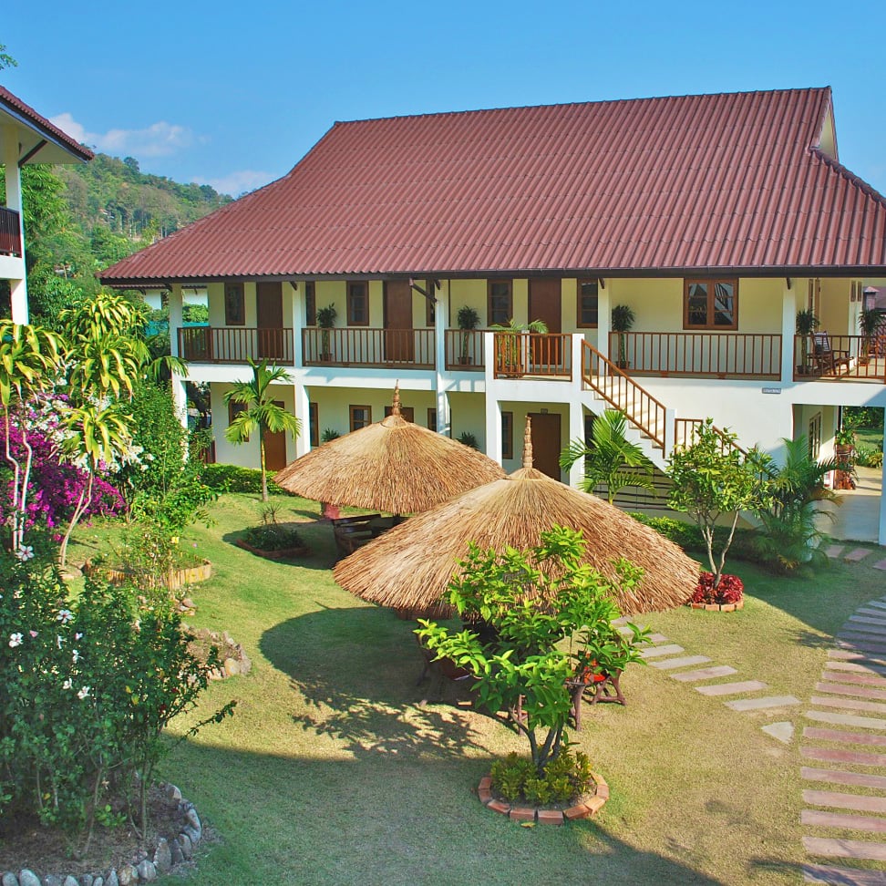 Resort courtyard with a two-story building featuring brown roof and wooden balconies, surrounded by a manicured lawn and tropical plants with thatched palapas in the foreground.