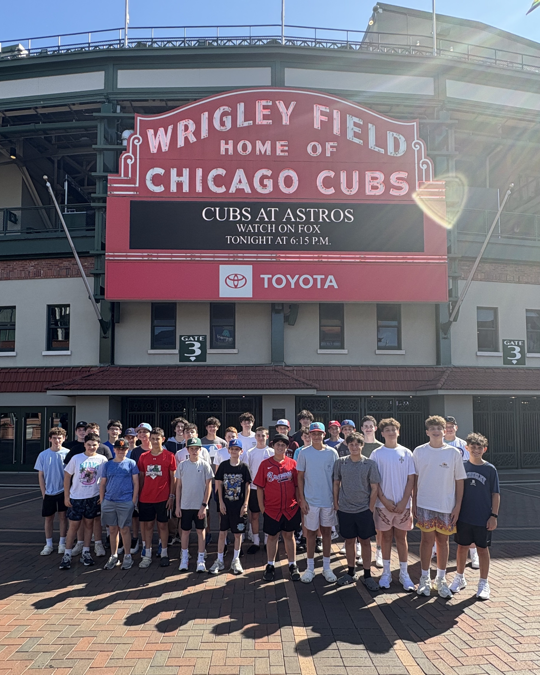 Group of young baseball players posing in front of Wrigley Field sign at the entrance, with a red marquee announcing Cubs games.