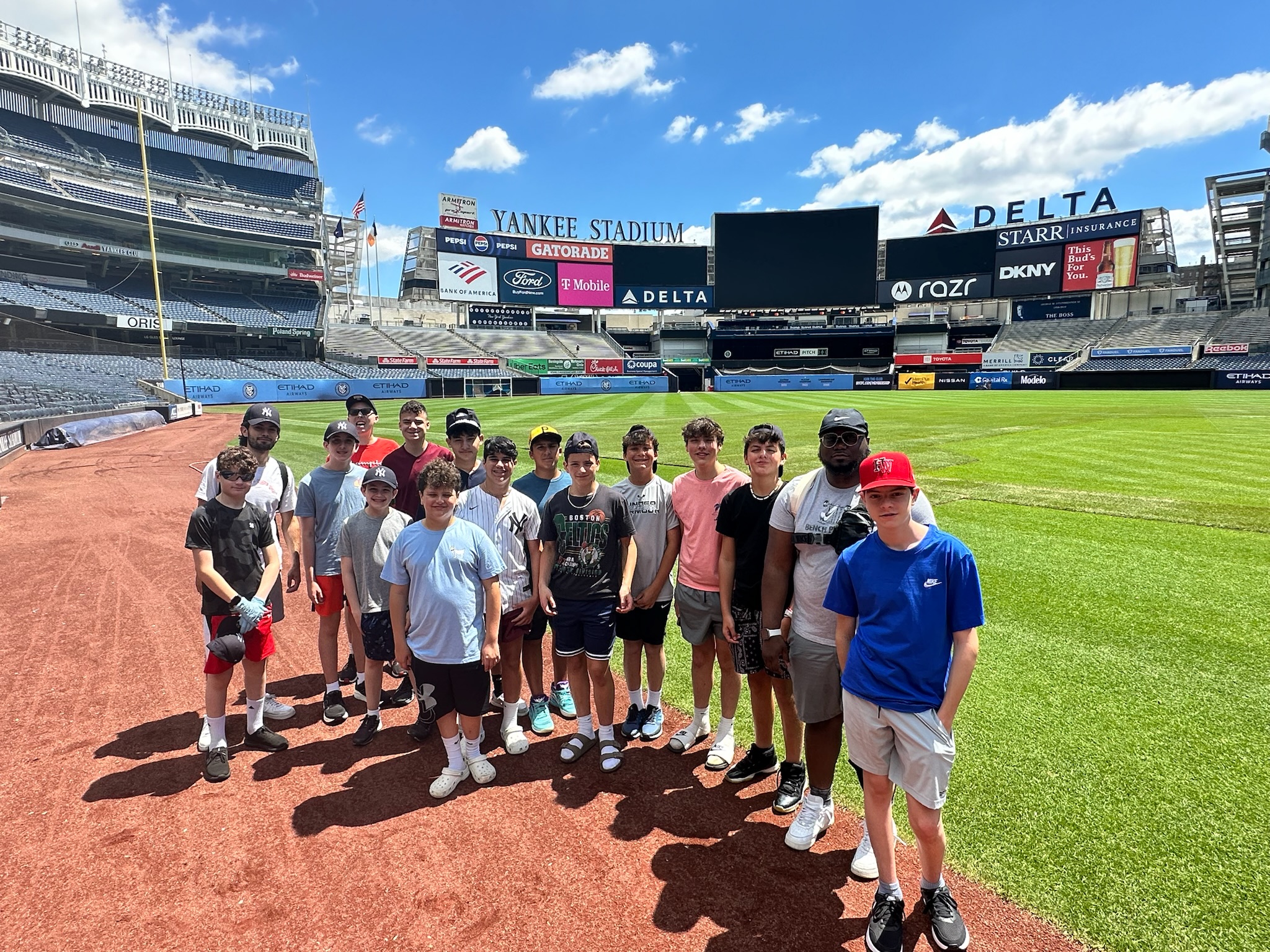 Group of kids posing for a photo on the infield at Yankee Stadium on a sunny day with stands in the background.