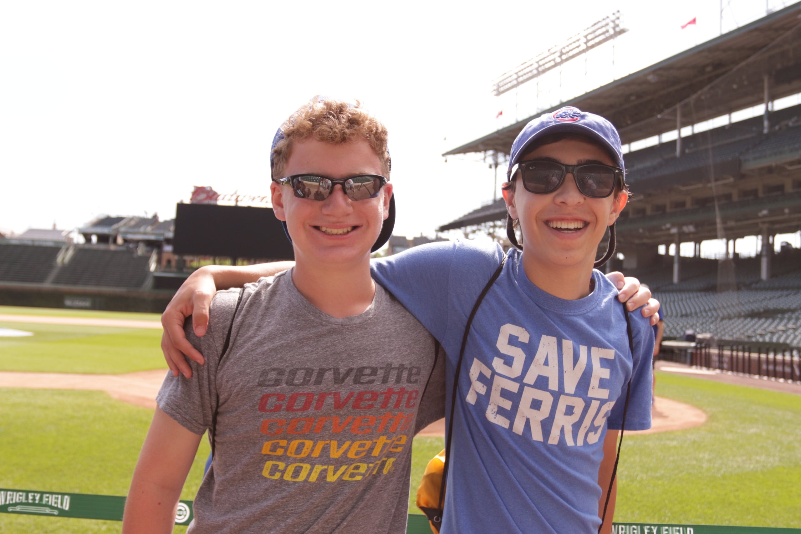 Two friends wearing sunglasses hug on a baseball field, with stands in the background and team shirts (gray Corvette top, blue shirt).